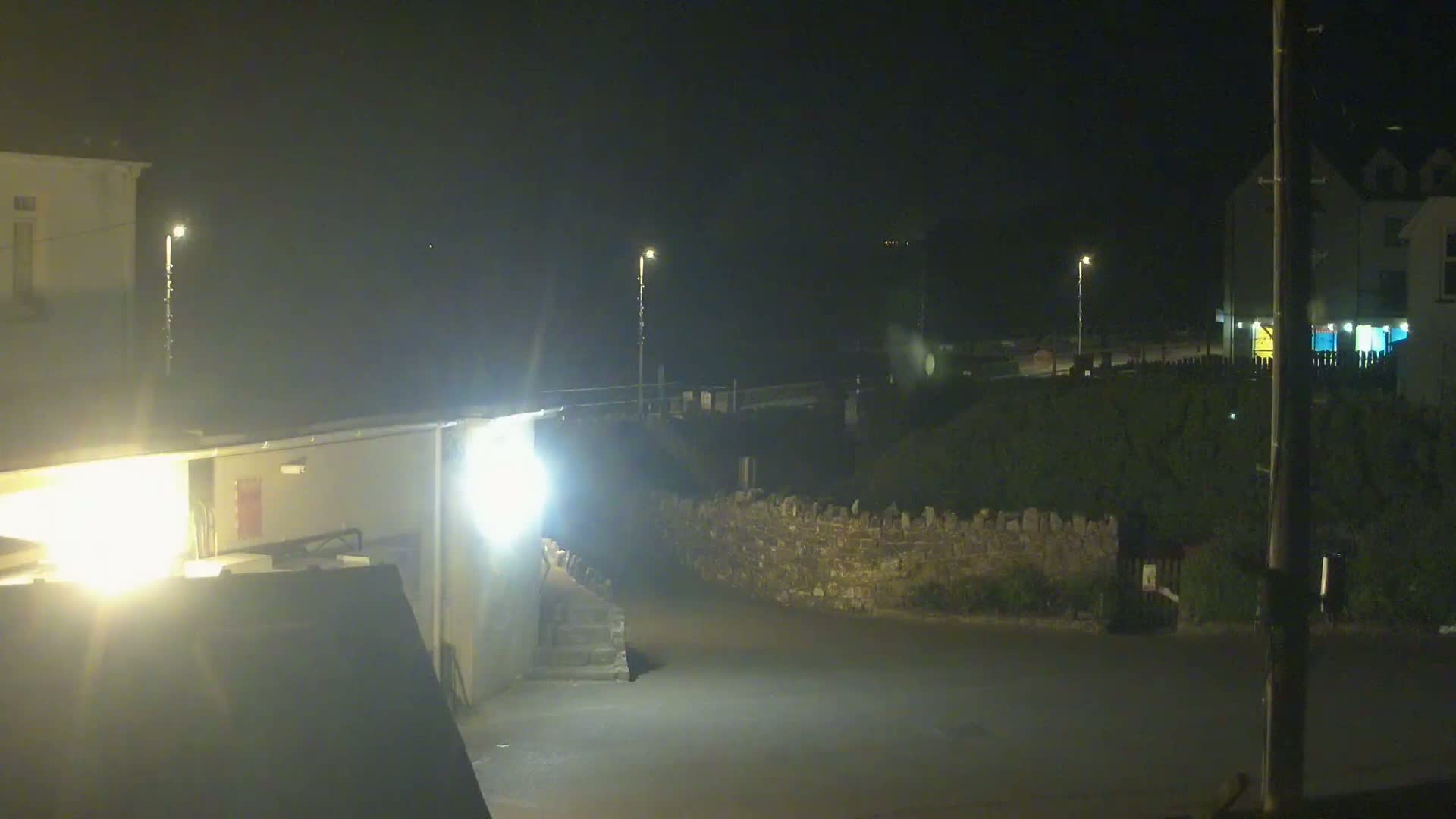 A nighttime street scene shows a low stone wall separating a paved area from some vegetation, with buildings and streetlights visible in the background under dark, clear skies.