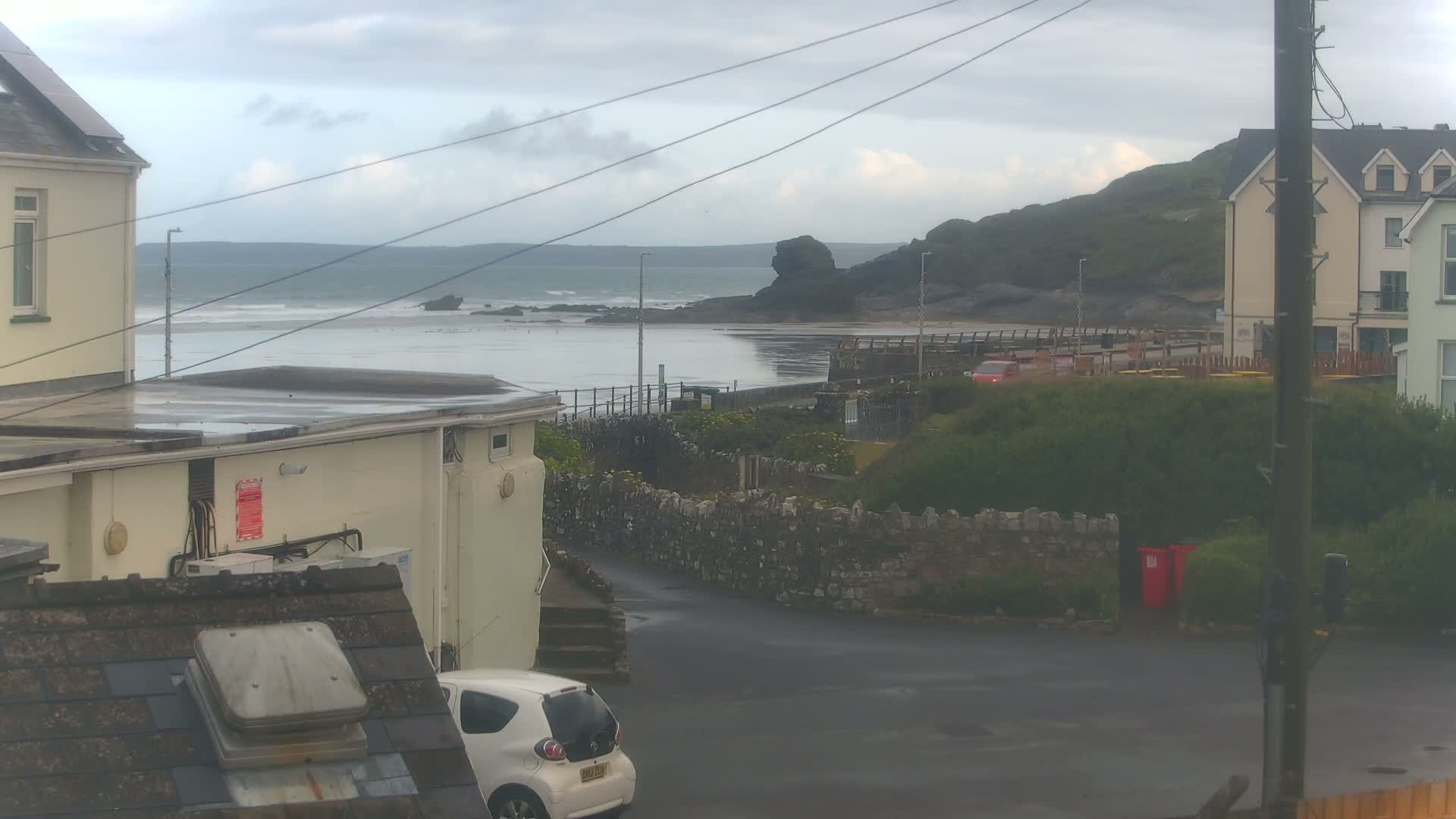 A wet, overcast day reveals a coastal scene with buildings, a small car, a low stone wall, and a beach with gentle waves.