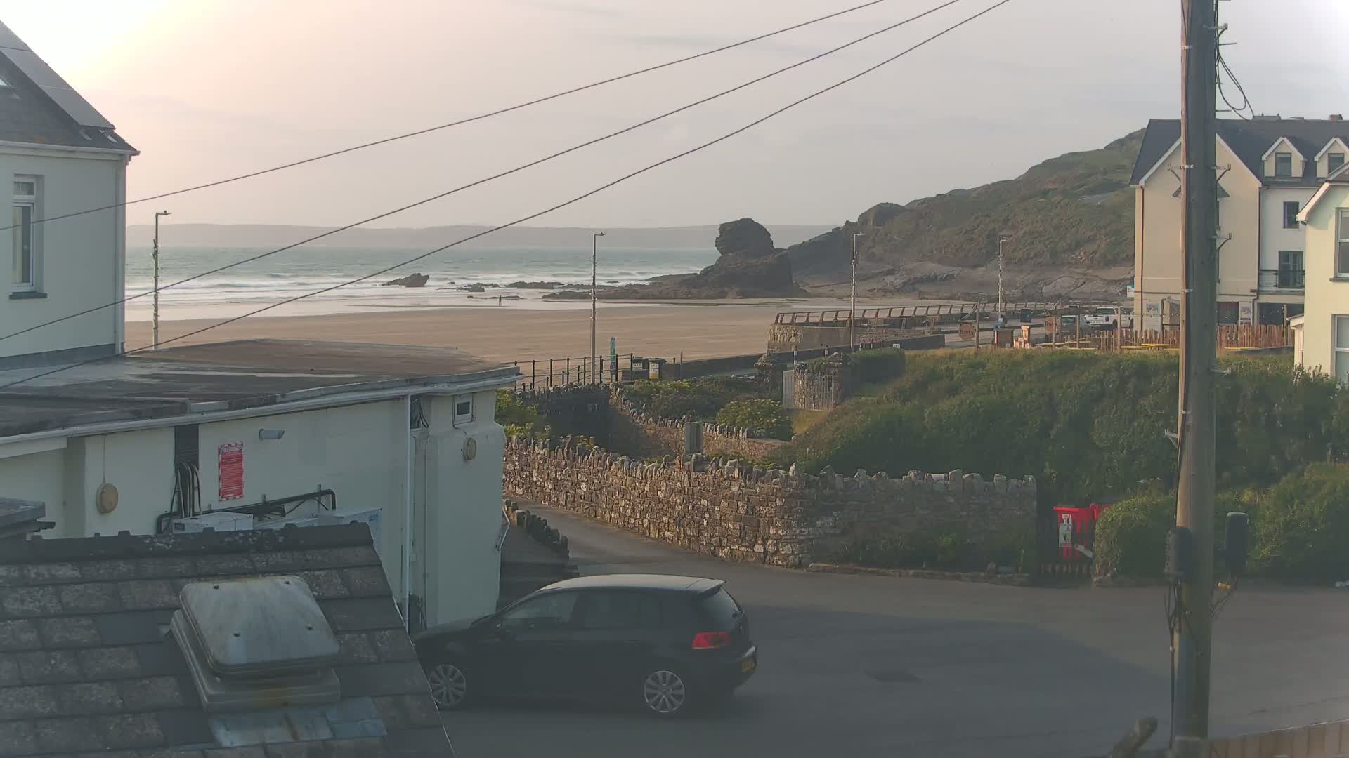 A sandy beach with gentle waves under a cloudy sky is viewed from between buildings, with a dark-colored car parked nearby.