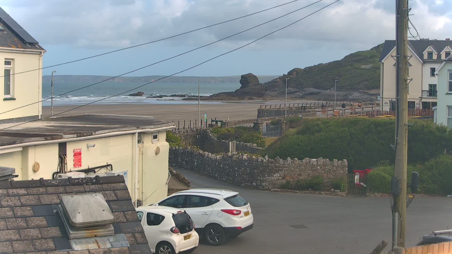 A view of a sandy beach and ocean under a cloudy sky, with buildings and two parked cars in the foreground.