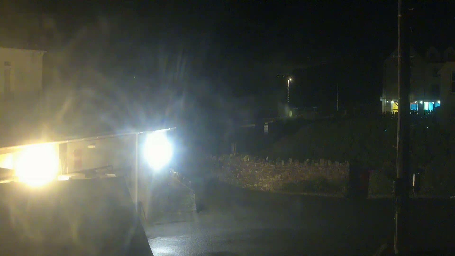 Headlights illuminate a wet, dark street at night, showing parts of buildings and a low stone wall.