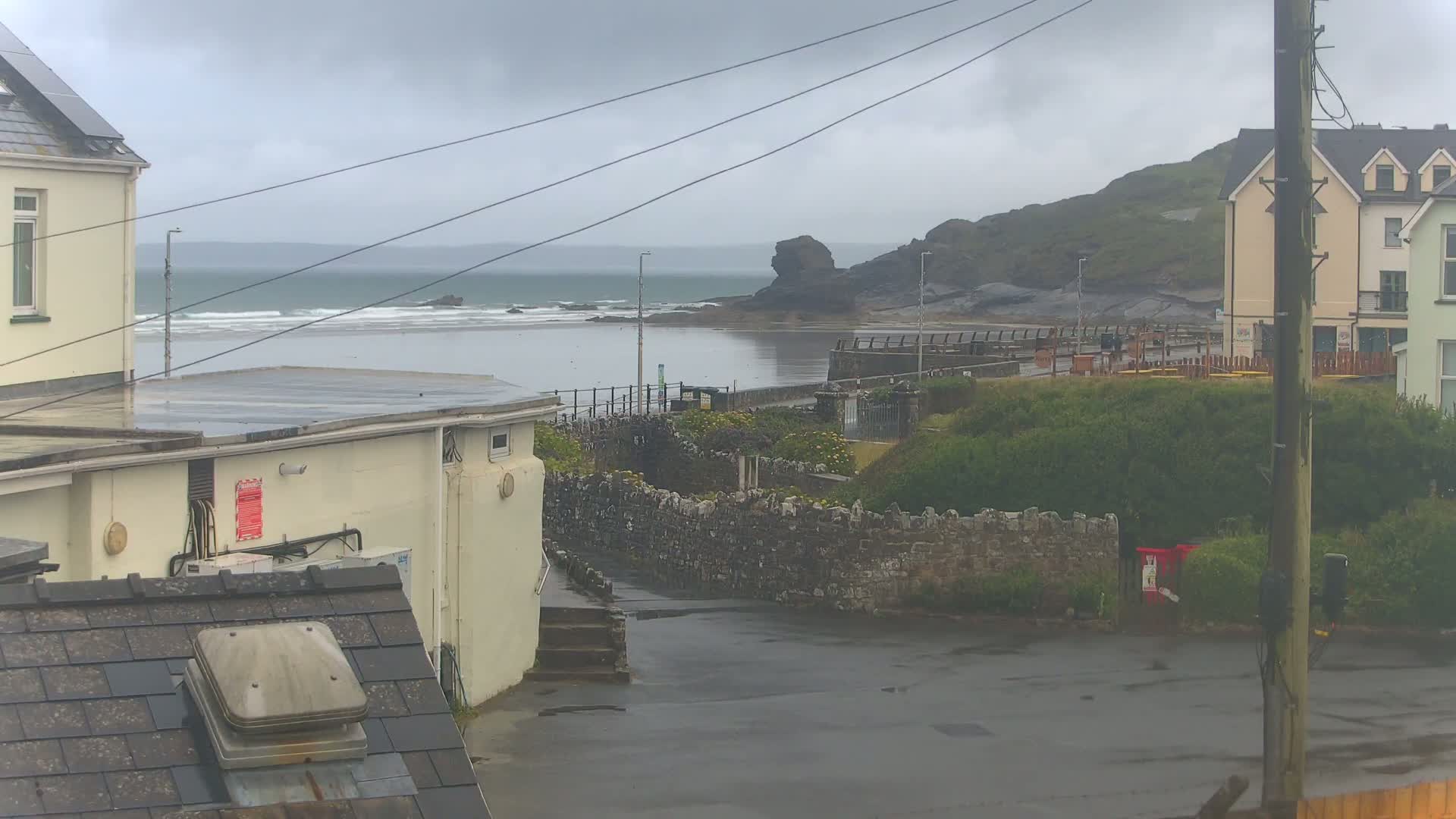 A wet coastal scene shows a gray, overcast sky, a calm sea with small waves, buildings, and a stone wall, all under a rainy sky.