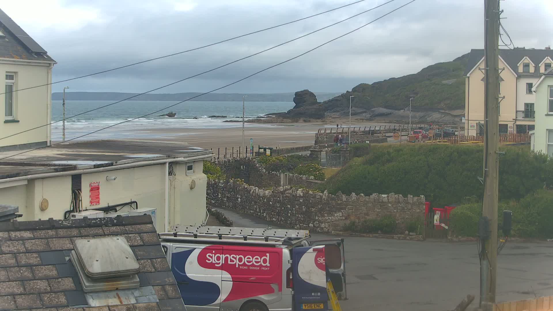 A view of a beach with moderate waves under an overcast sky, seen from between two buildings, with a van parked in the foreground.