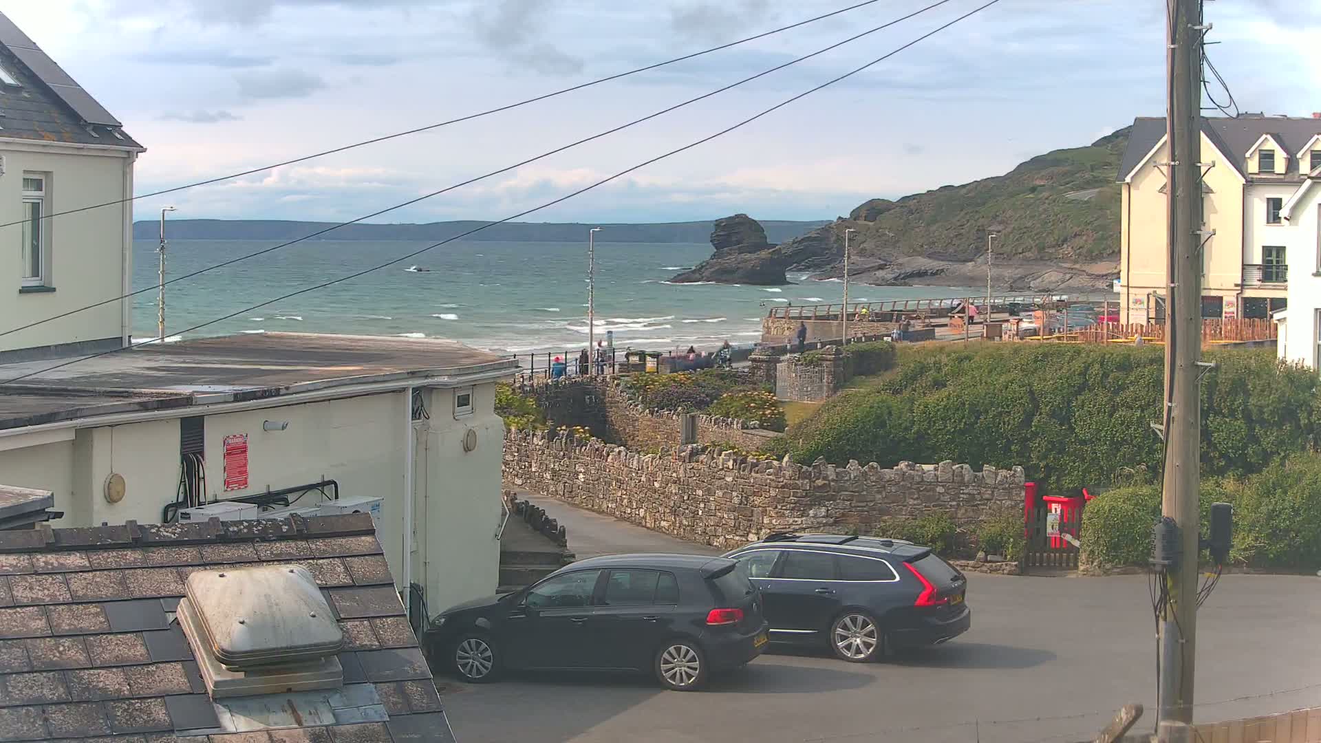 Two dark-colored cars are parked on a street overlooking a rocky coastline with a wavy ocean under a partly cloudy sky.