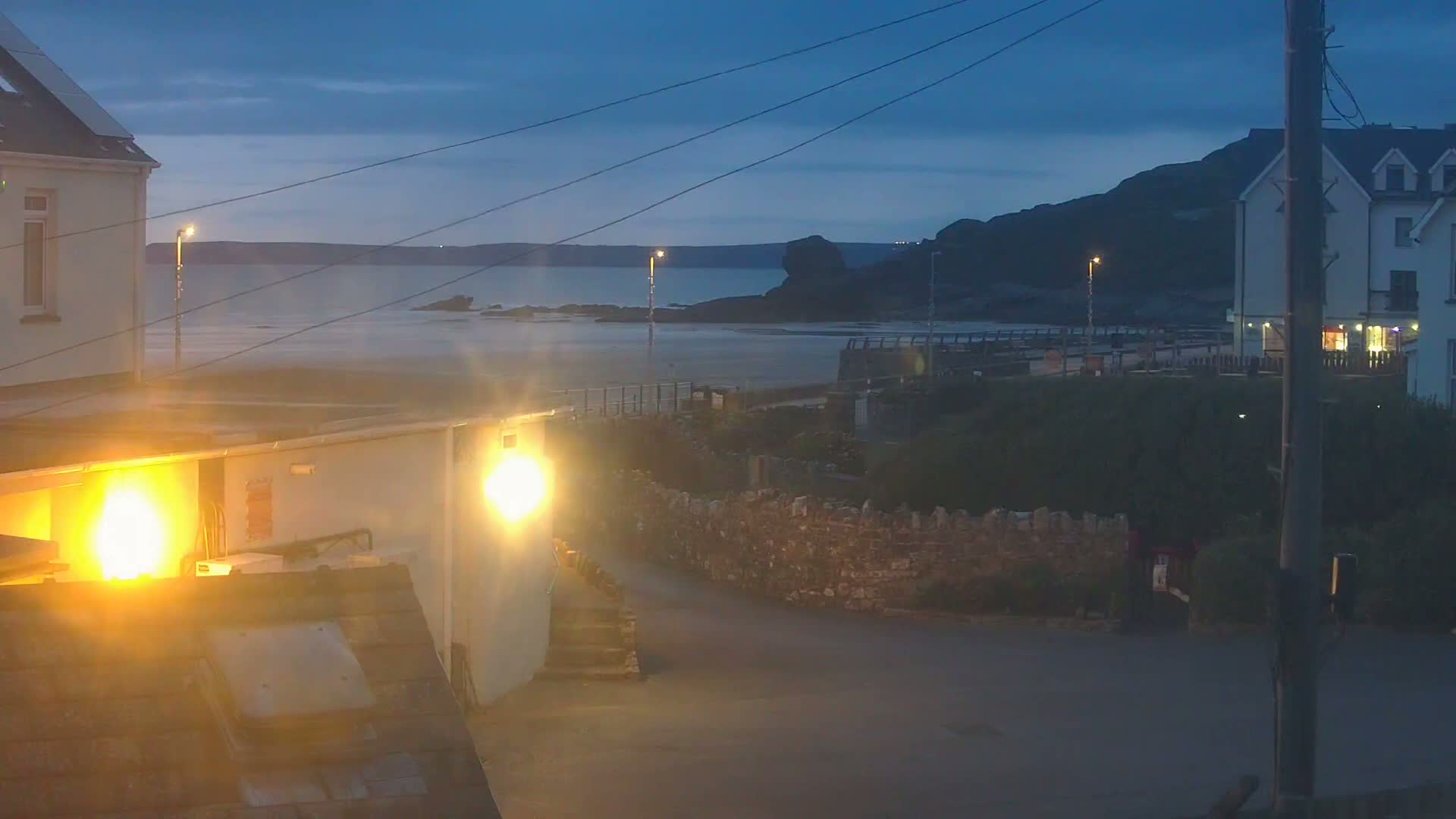 A dark, twilight view of a beach and coastal buildings, with low-lying lights illuminating a small street.