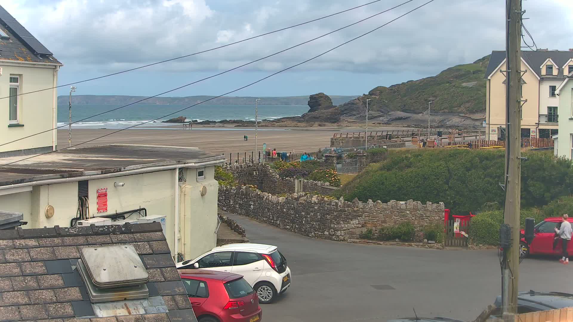 A view from a rooftop shows a sandy beach with a few people and some waves under an overcast sky, with parked cars and buildings in the foreground.