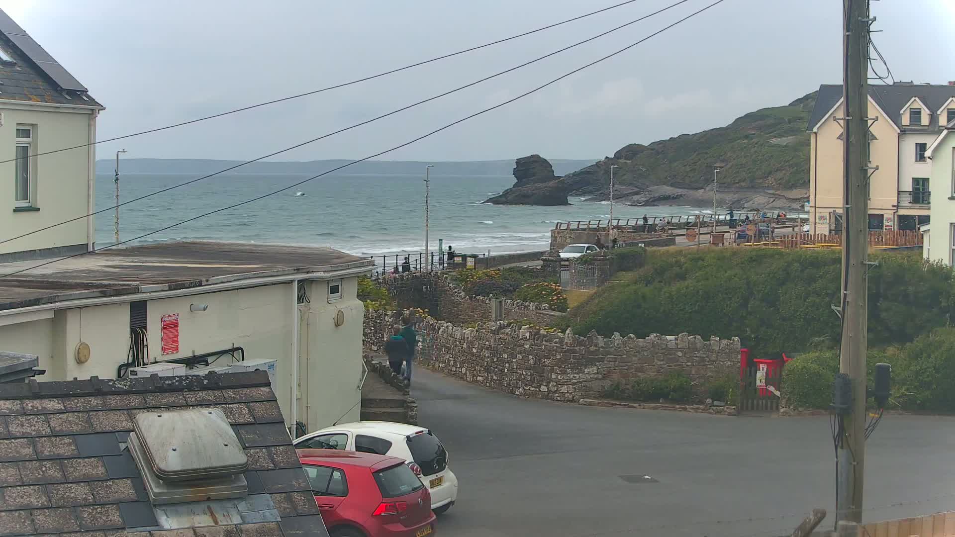 A view of a rocky coastline with a choppy sea under an overcast sky, seen from a residential area with parked cars and buildings.