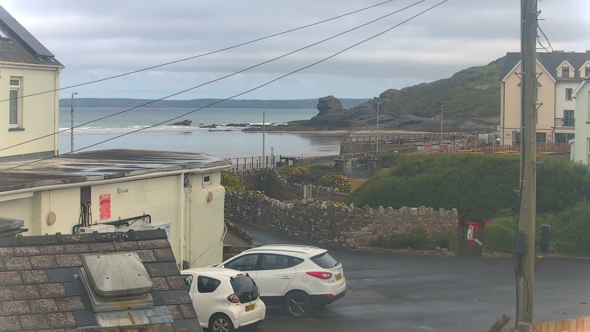 Two white cars are parked in a coastal town's parking lot, overlooking a calm ocean under an overcast sky.