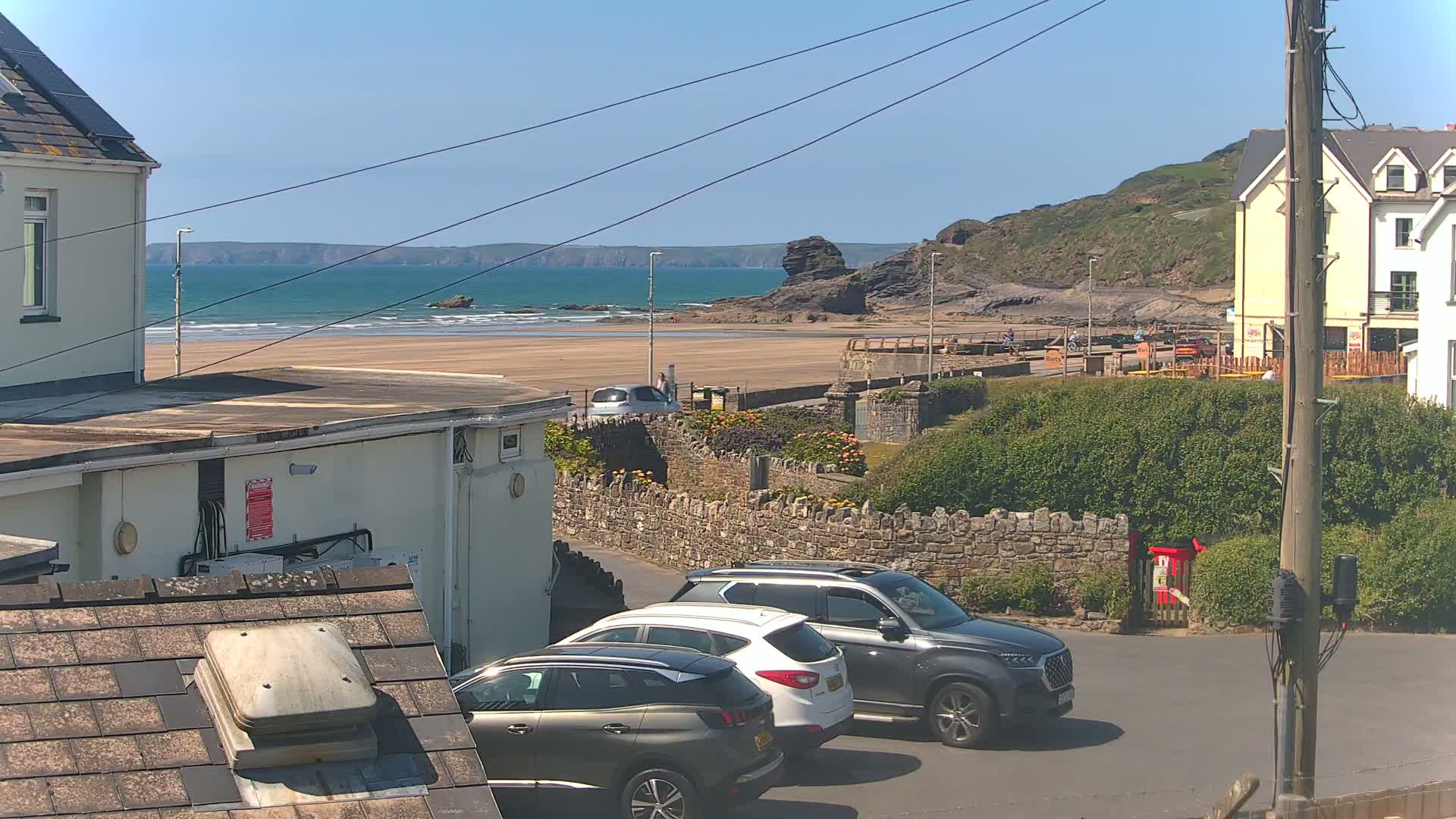 A sunny day overlooking a sandy beach with calm ocean waves, several parked cars, and houses nestled on a hillside.