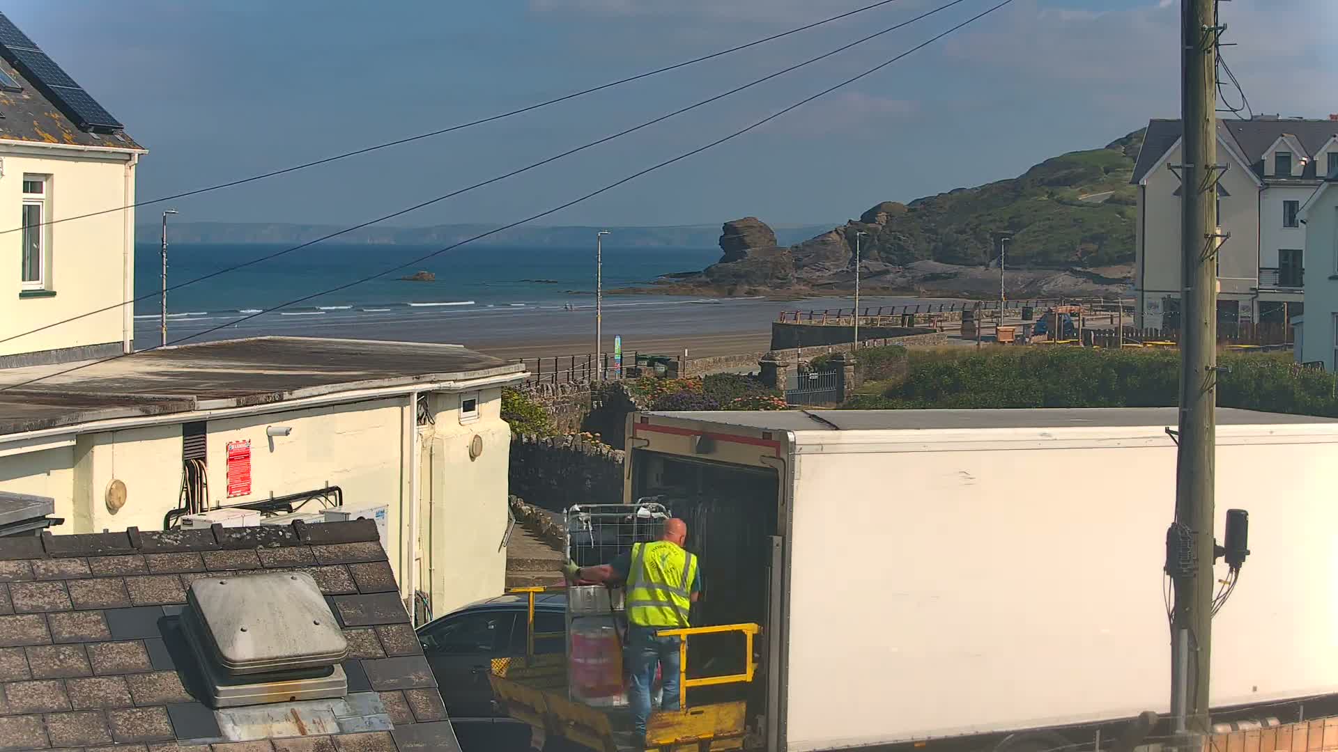 A man in a yellow vest loads a shopping cart into a white delivery truck near a beach on a sunny day.