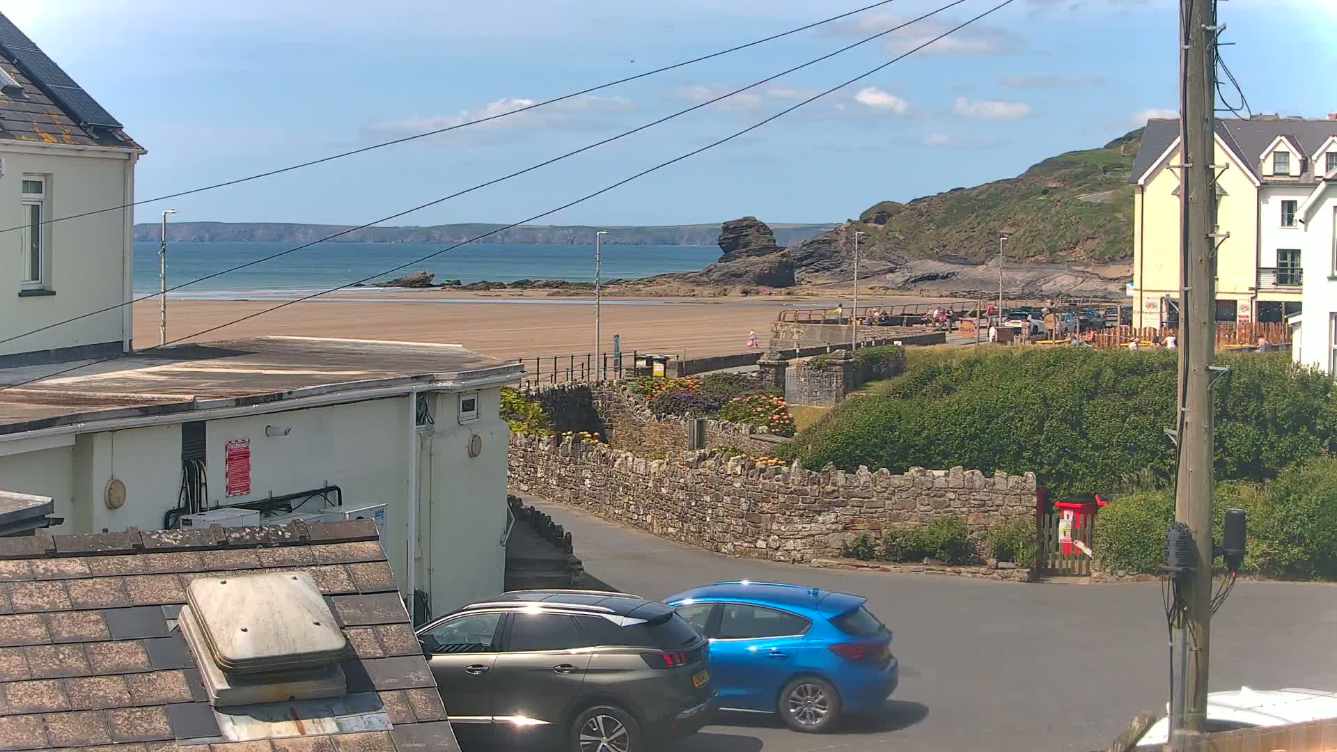 A sandy beach with a calm ocean is visible beyond a stone wall and parked cars, under a mostly sunny sky.
