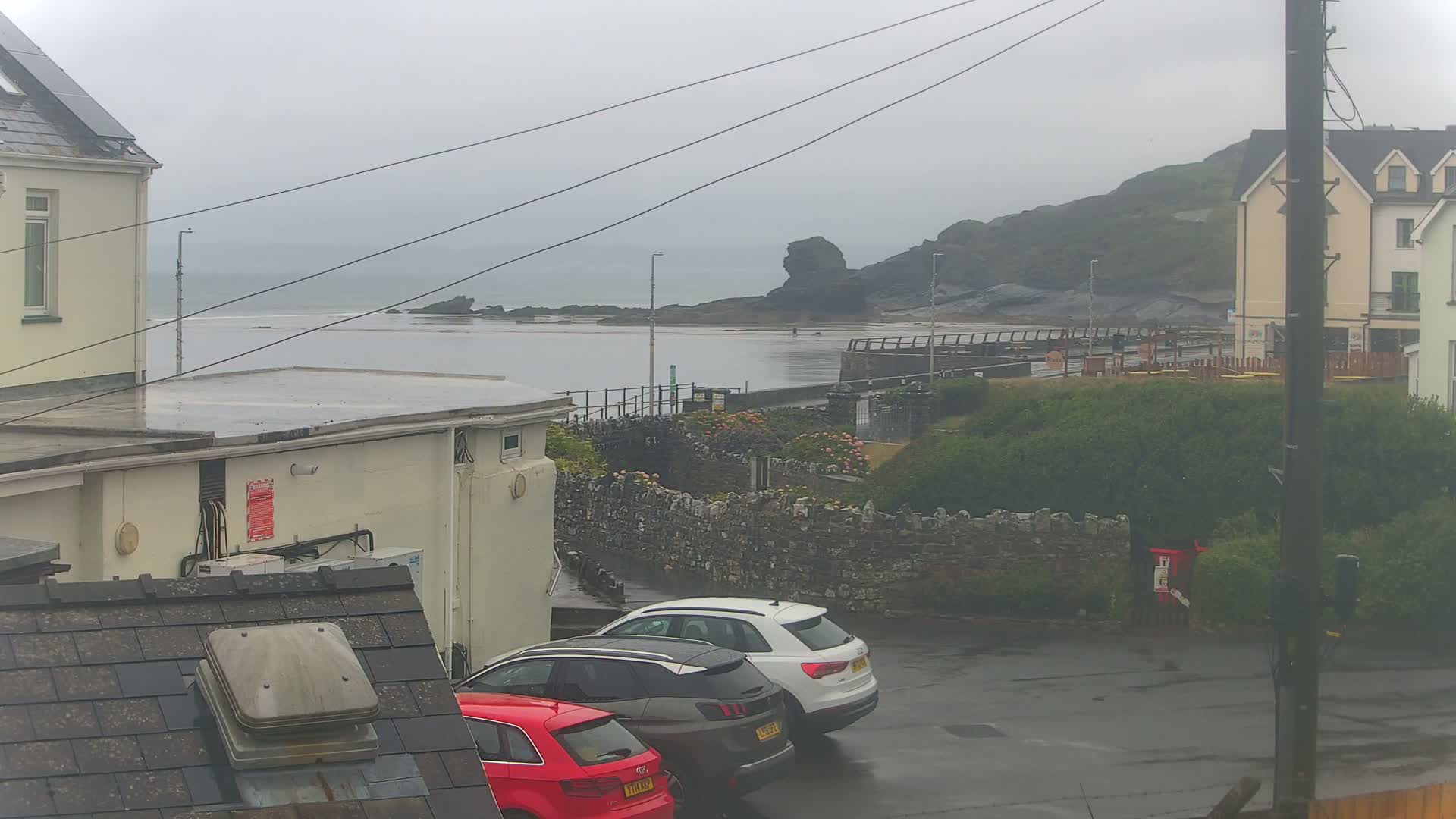A wet, overcast day reveals a coastal scene with parked cars in a lot overlooking a calm sea and rocky shoreline, partially obscured by buildings and vegetation.