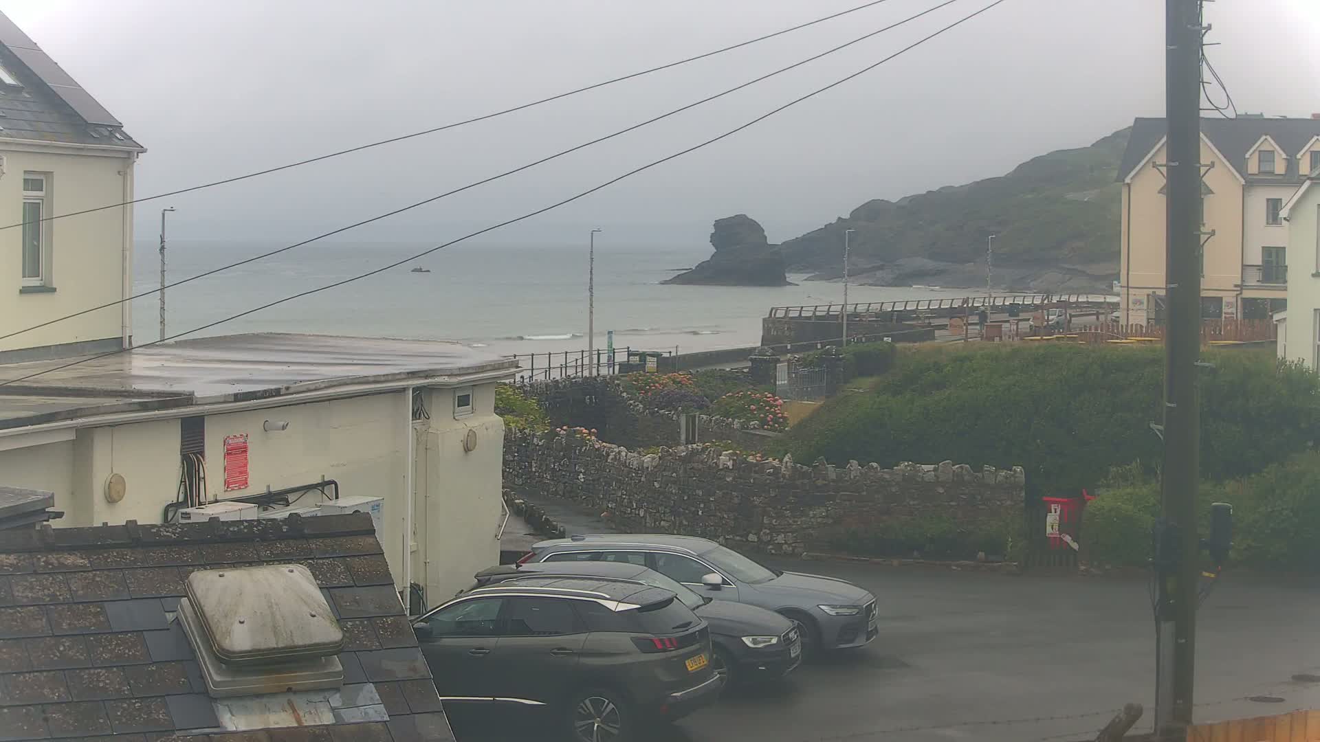 A rainy day overlooks a coastal town with parked cars, a stone wall, and a distant rocky outcrop visible through an overcast sky.