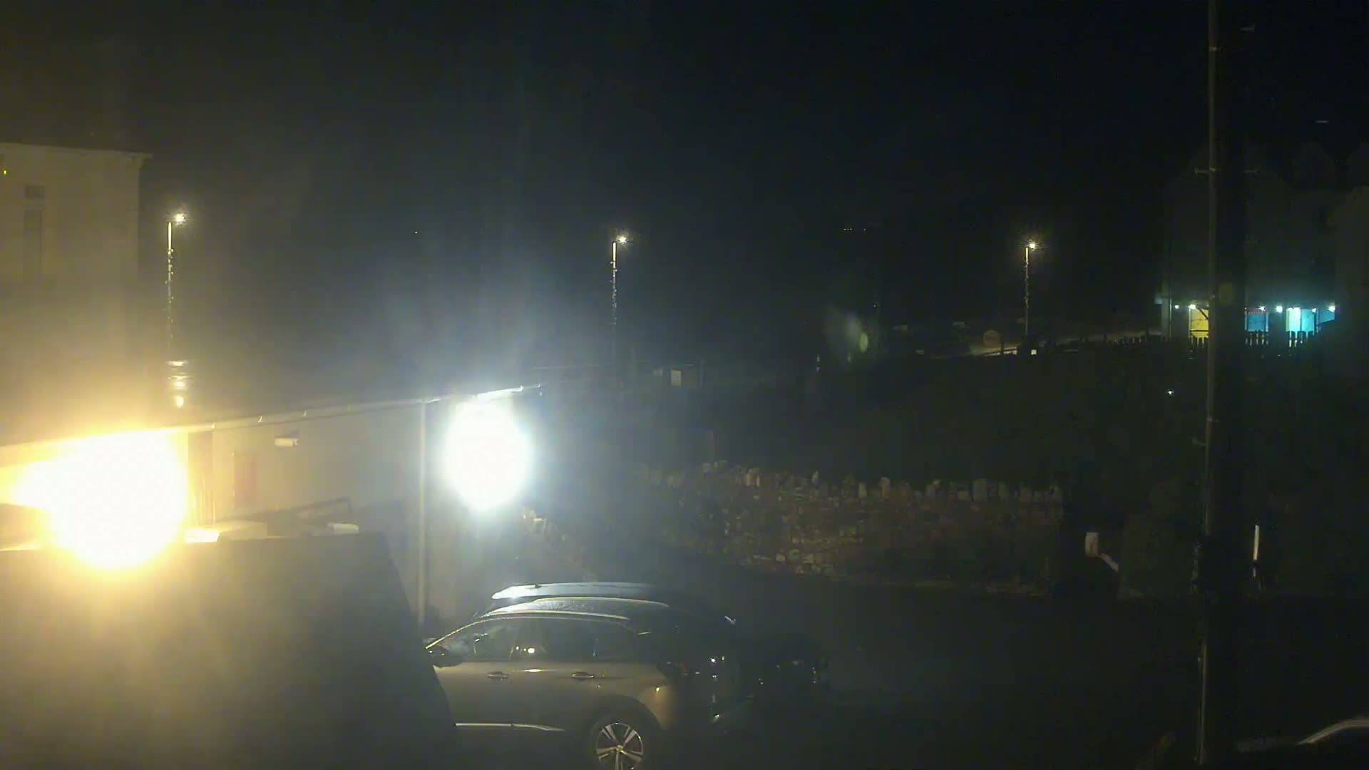 A nighttime, possibly rainy, scene shows a silver SUV parked in a dimly lit area near buildings and a low wall.