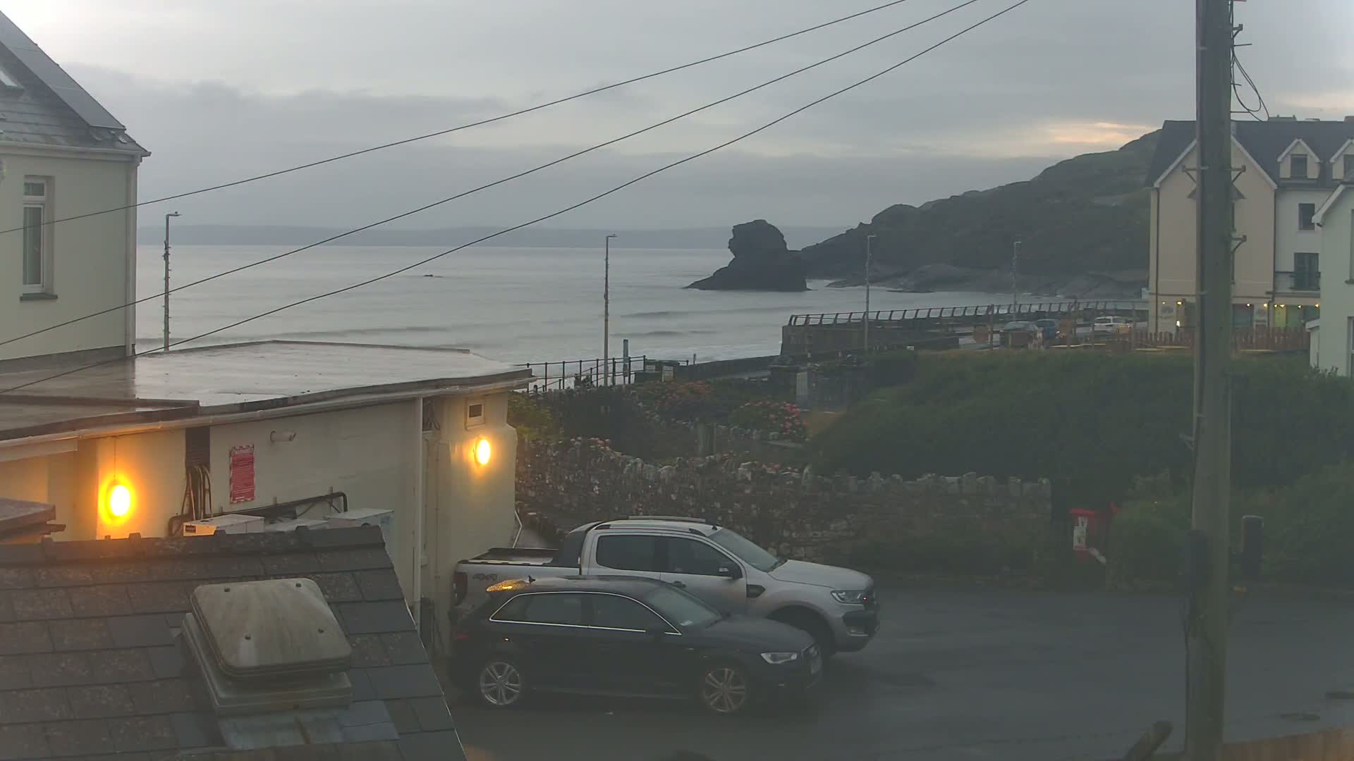 A wet, overcast day shows a coastal scene with two cars parked near buildings, overlooking a calm sea and a rocky outcrop.