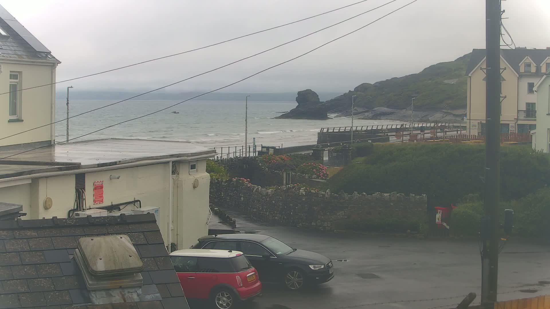 A wet, overcast day shows a coastal scene with two cars parked near buildings, overlooking a calm sea and a rocky outcrop.