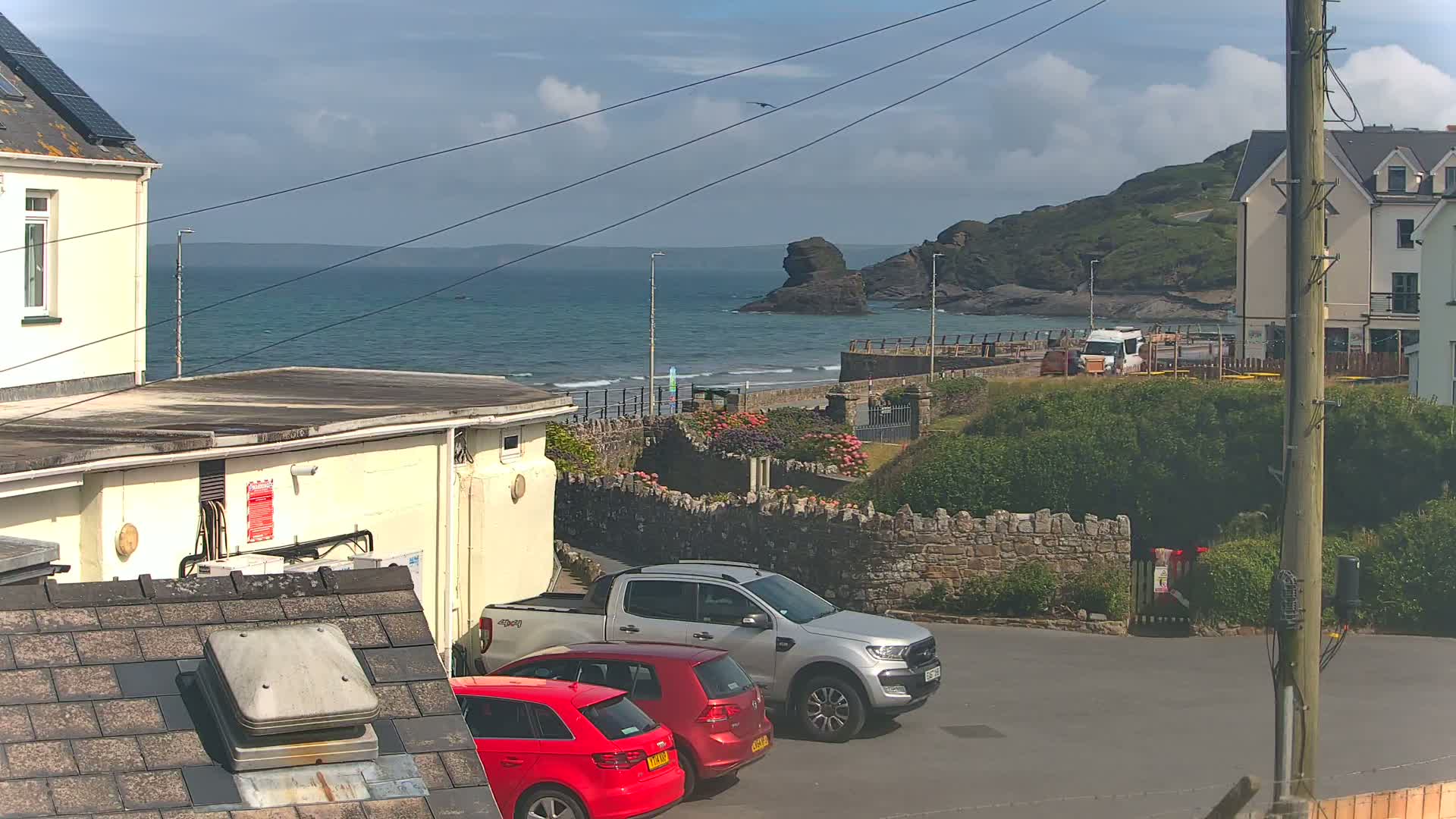 A partly sunny day overlooks a coastal scene with a calm ocean, a sandy beach, buildings, and parked cars, including a silver pickup truck and two red cars.