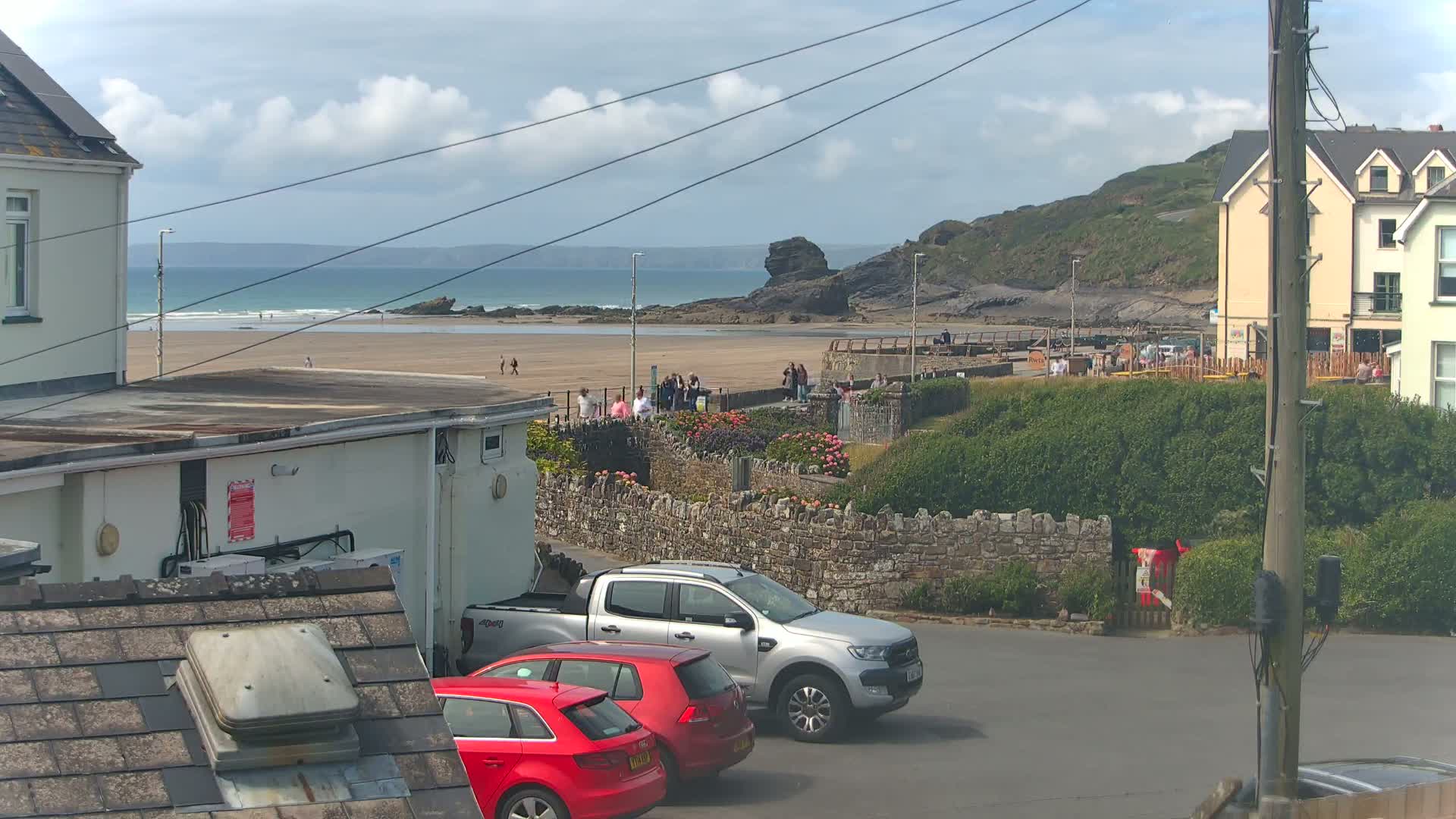 A partly cloudy day overlooking a sandy beach with a calm ocean, people walking along a stone wall, and parked cars near buildings.