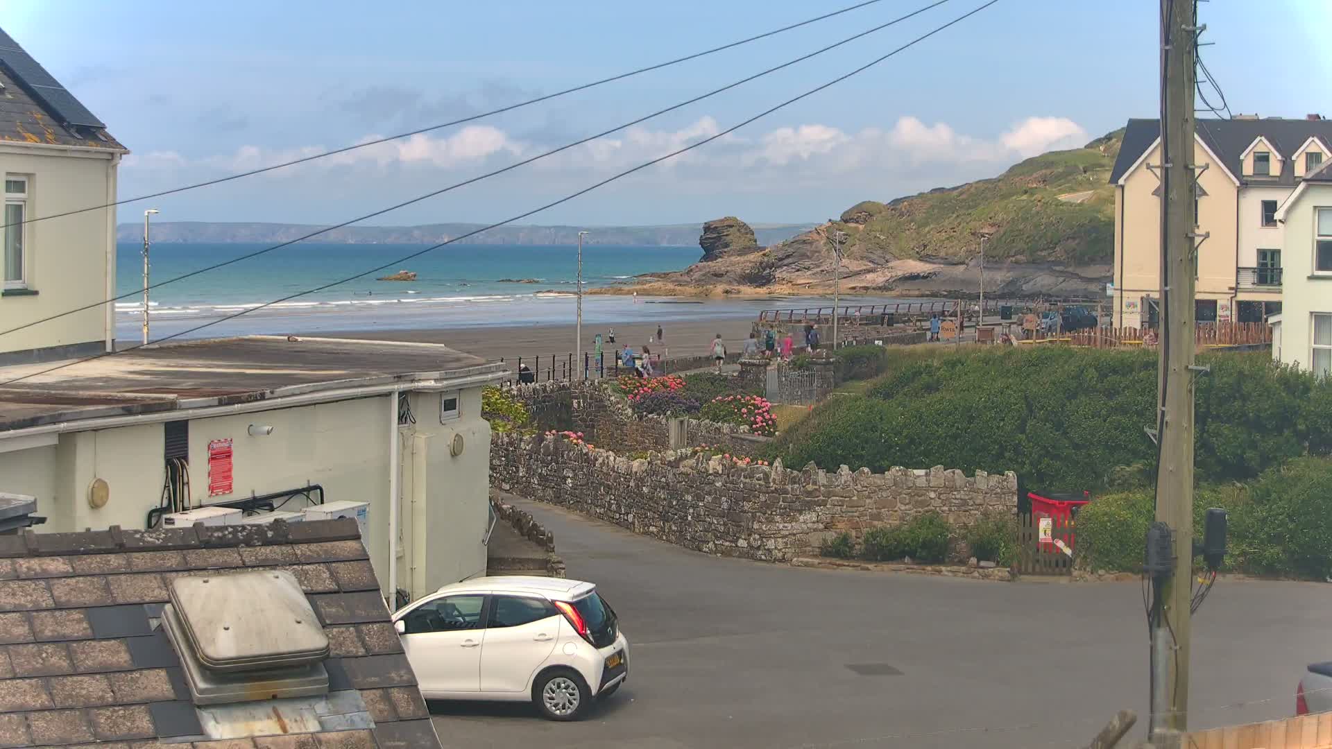 A view of a sandy beach with people walking along the shore under a partly cloudy sky, seen from a residential area with a white car parked on a street next to a stone wall.