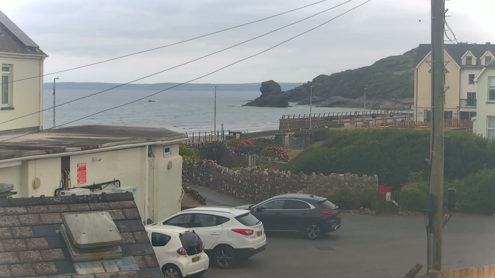 A view from a rooftop shows a calm, overcast day at the coast, with two parked cars in a small parking area next to buildings overlooking the sea and a rocky outcropping.