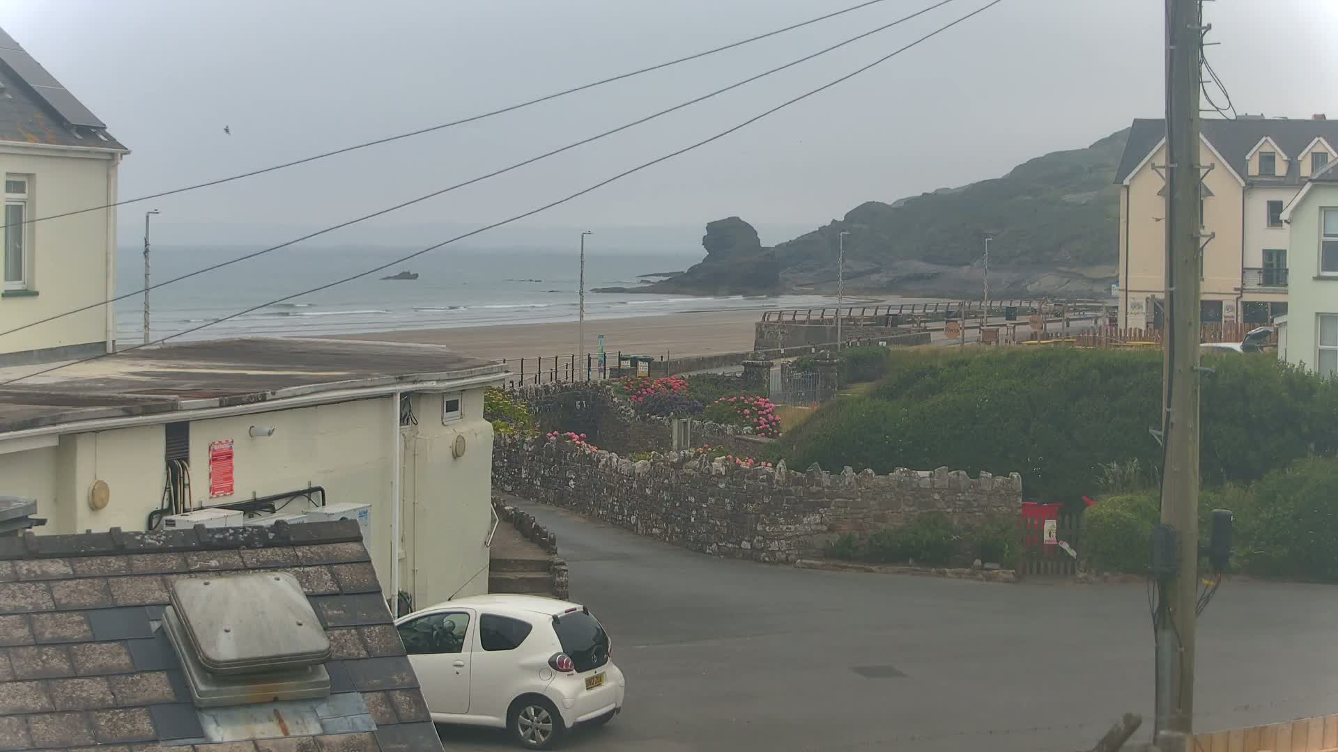 A view of a beach with a sandy shore and gentle waves under an overcast sky, seen from a residential area with houses and a small white car parked on a street.