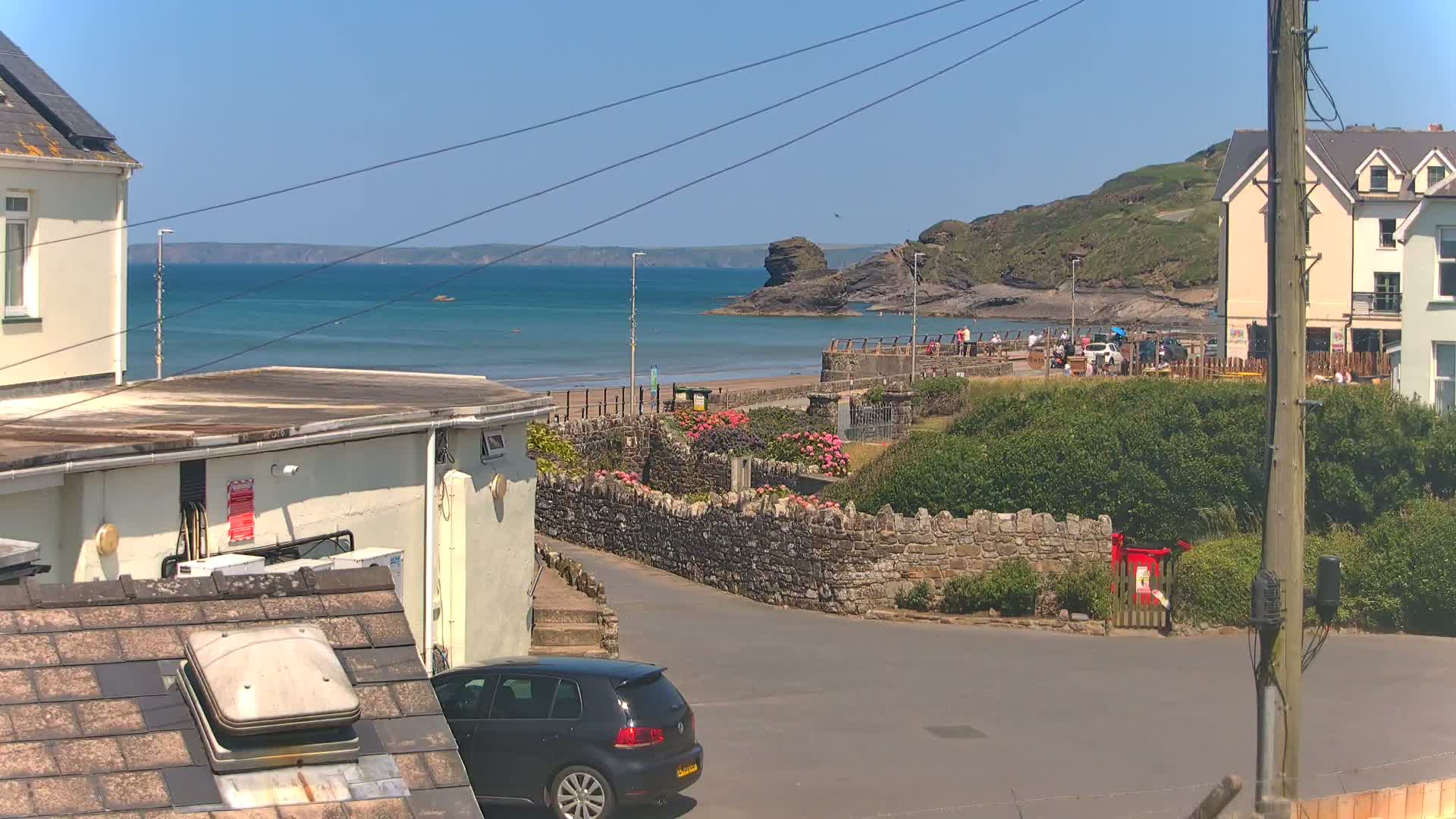 A sunny seaside view shows a calm ocean, a sandy beach with people, stone walls, buildings, and a dark-colored car parked near a street.