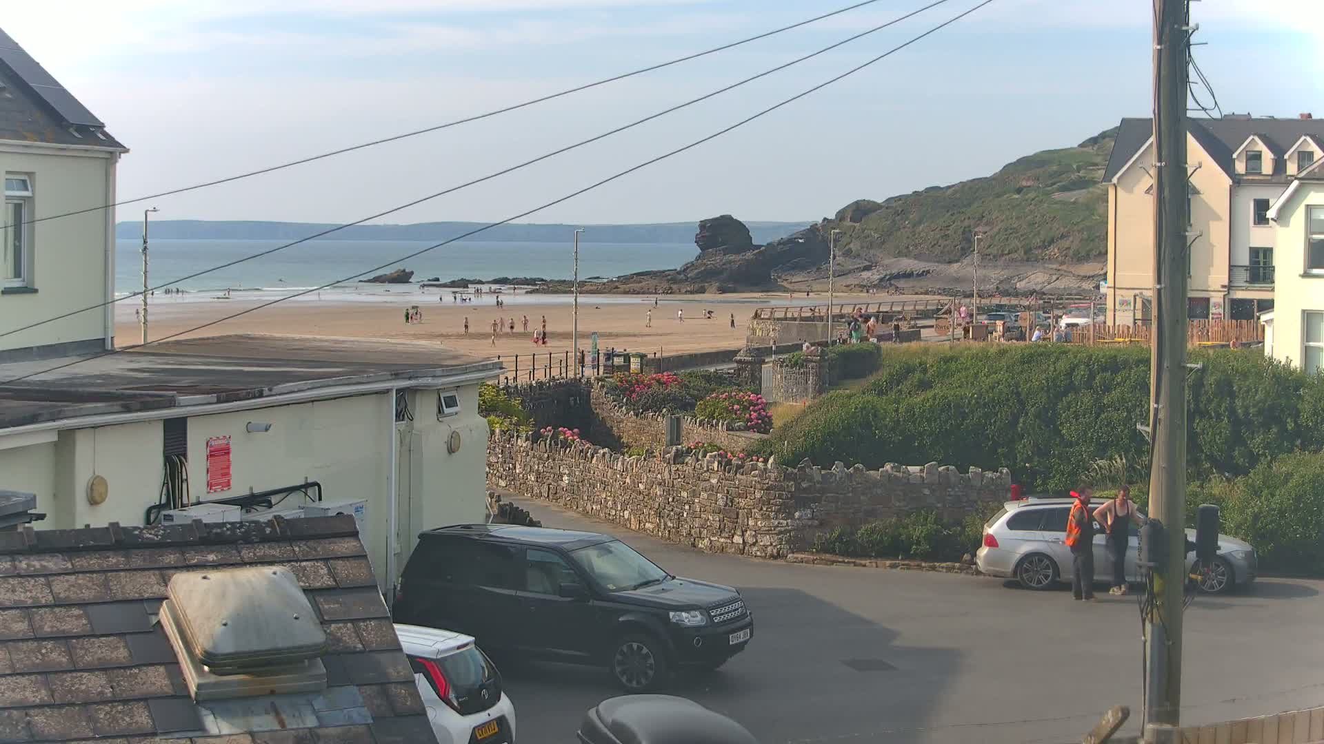A sandy beach with people on it is visible from a residential area on a partly sunny day, with cars parked on a street in the foreground.