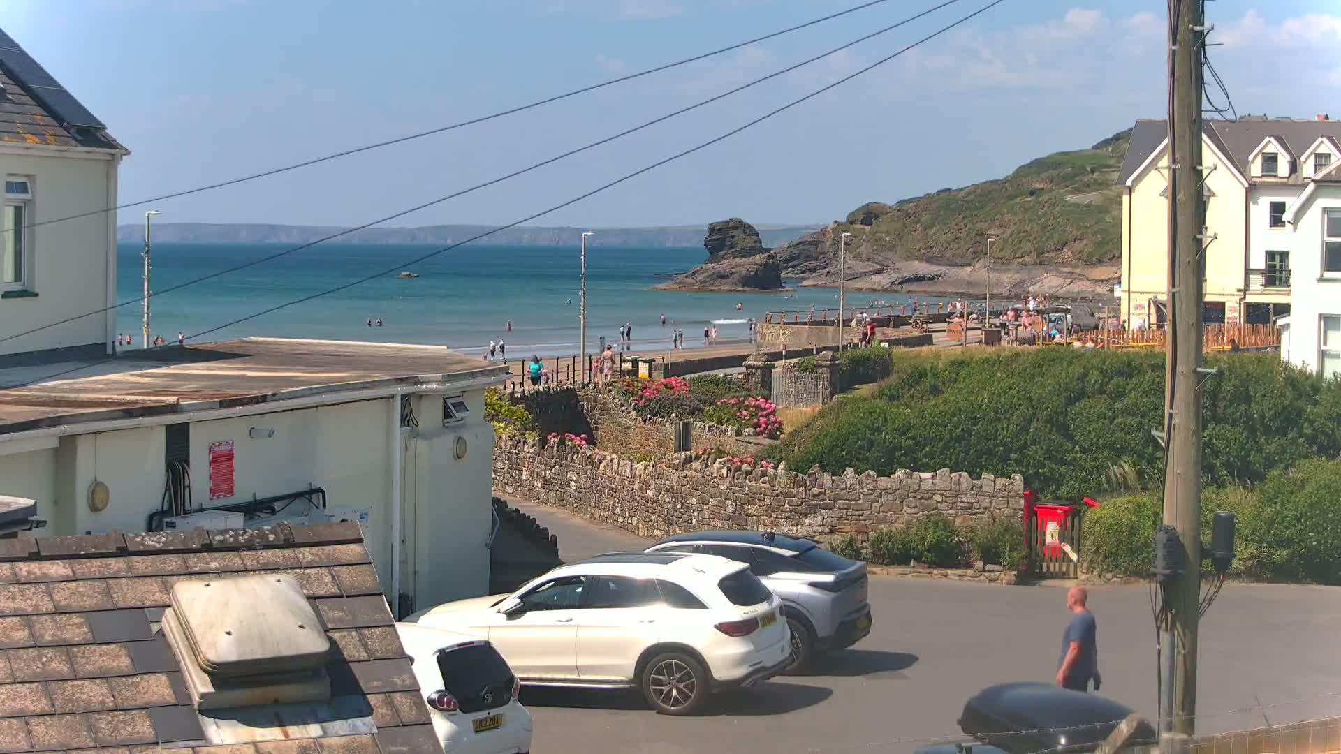 A sunny day at a seaside town shows a beach with people swimming and sunbathing, two cars parked on a street near buildings, and a man walking by a red post box.