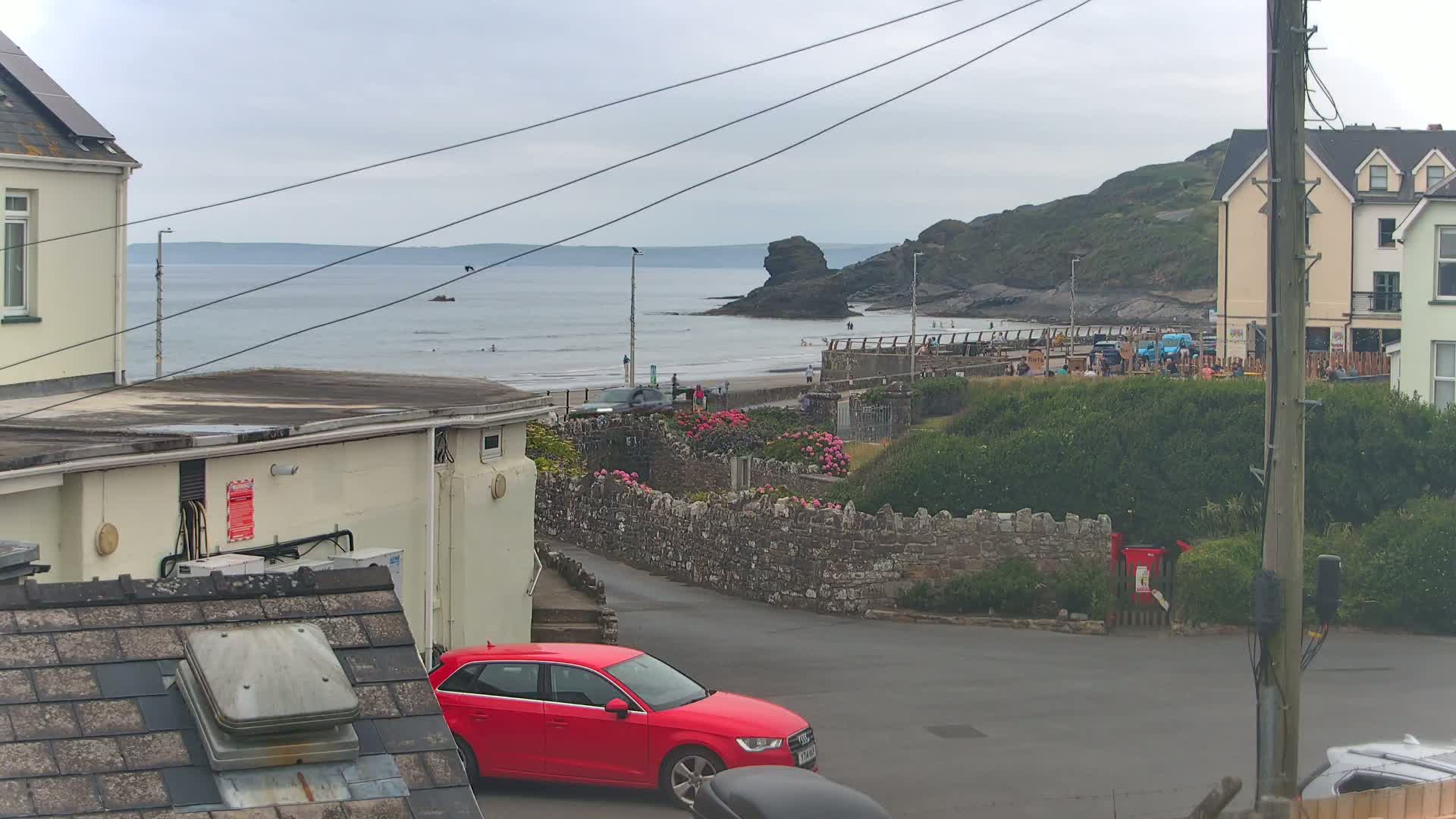A red car is parked near a stone wall overlooking a beach with people on it, a calm sea, and a cloudy sky.