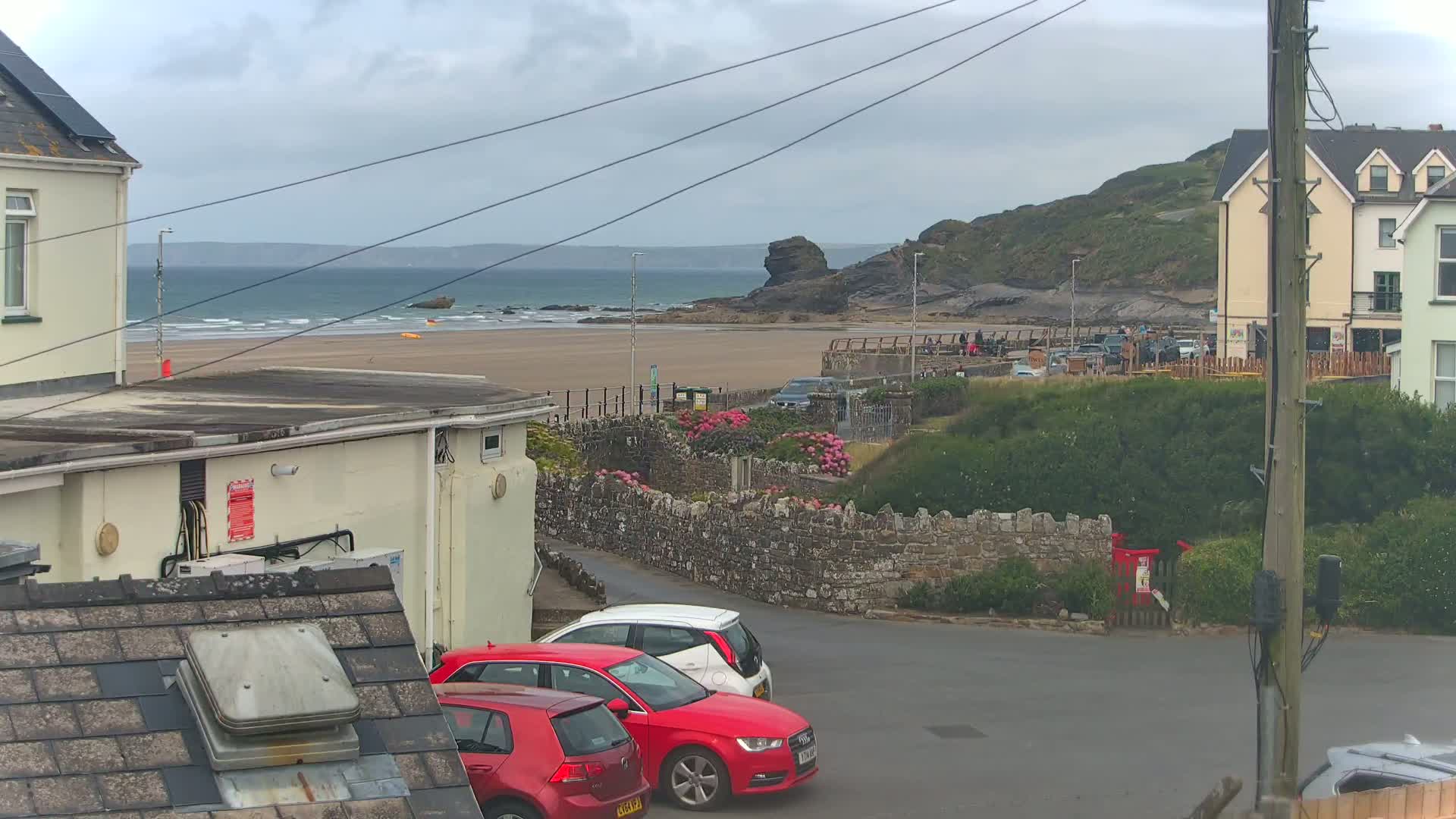 A sandy beach with a calm ocean stretches behind a row of parked cars near buildings, under an overcast sky.