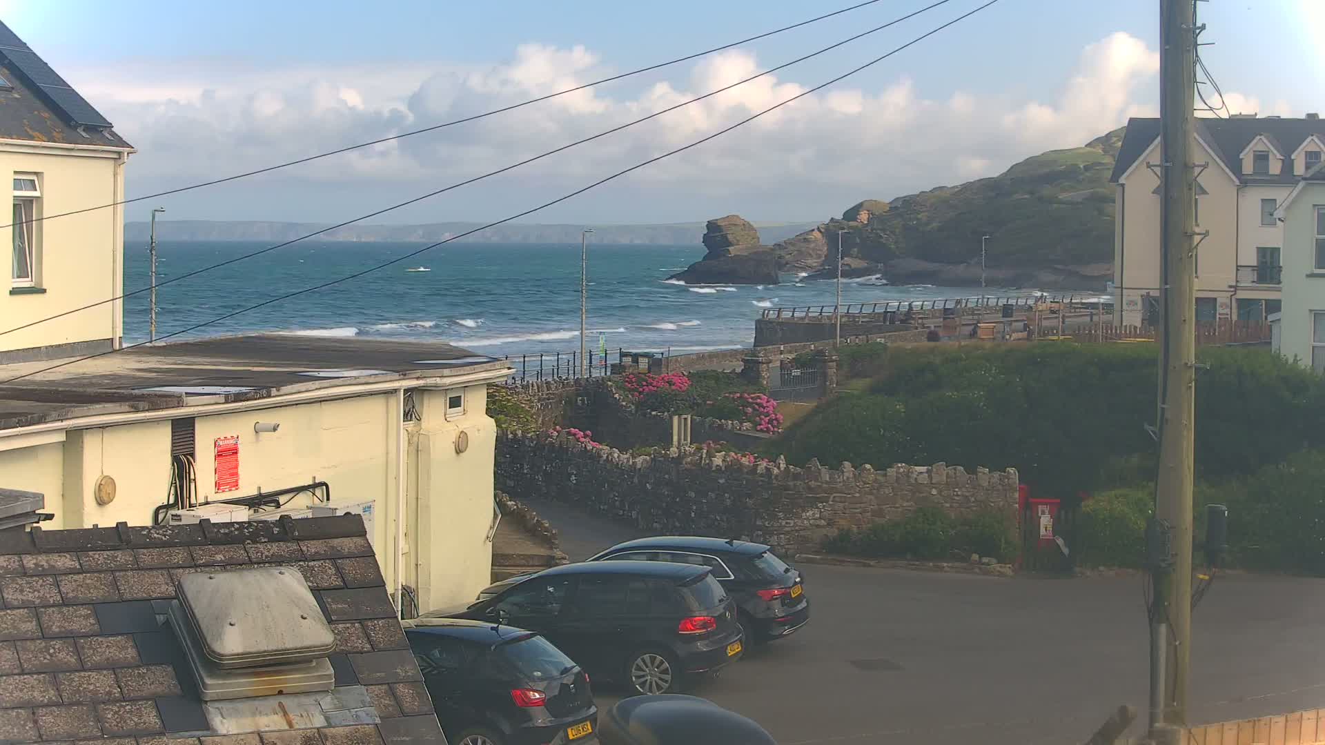 A view of a rocky coastline with a somewhat choppy sea under a partly cloudy sky, seen from a parking area between buildings with several parked cars.