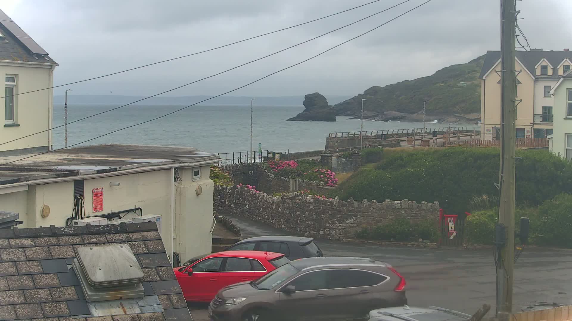 A cloudy day overlooks a coastal scene with parked cars, buildings, and a rocky outcrop in the sea.