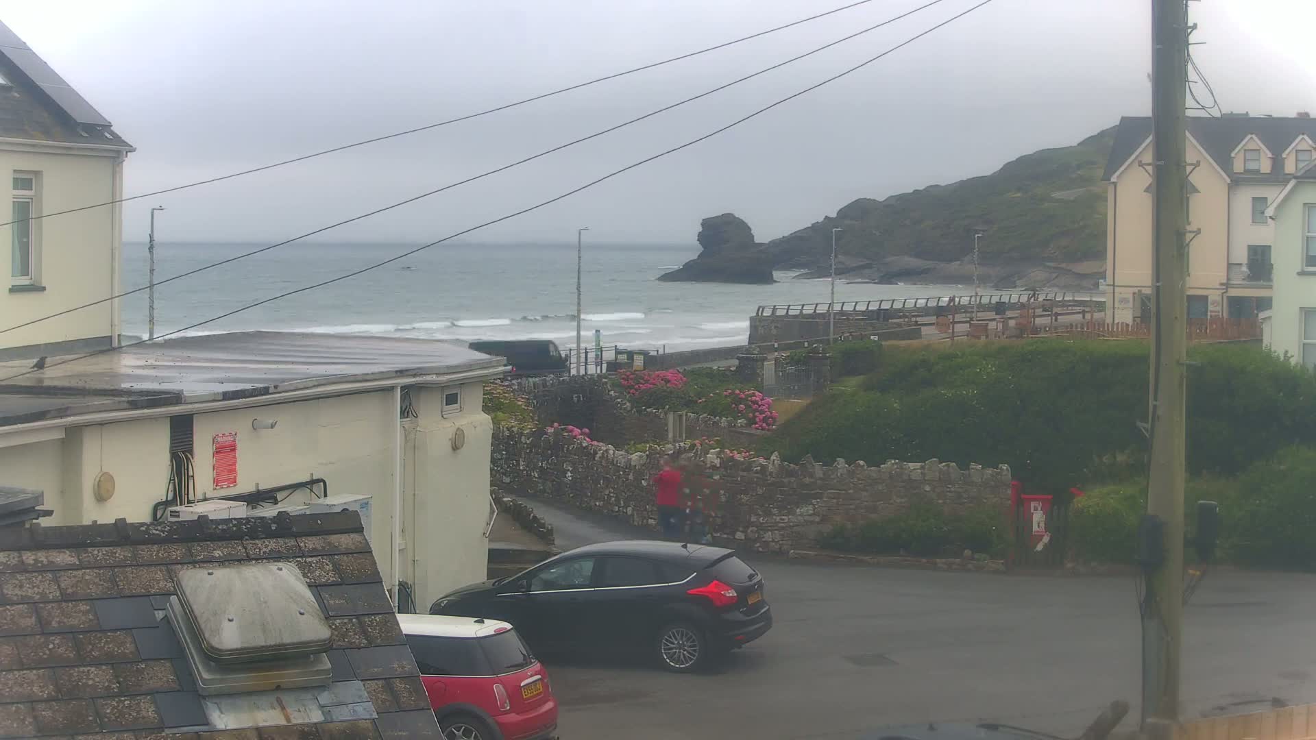 A coastal scene on an overcast day shows a gray sea with small waves, buildings, cars parked on a street, and a few people walking near a stone wall.