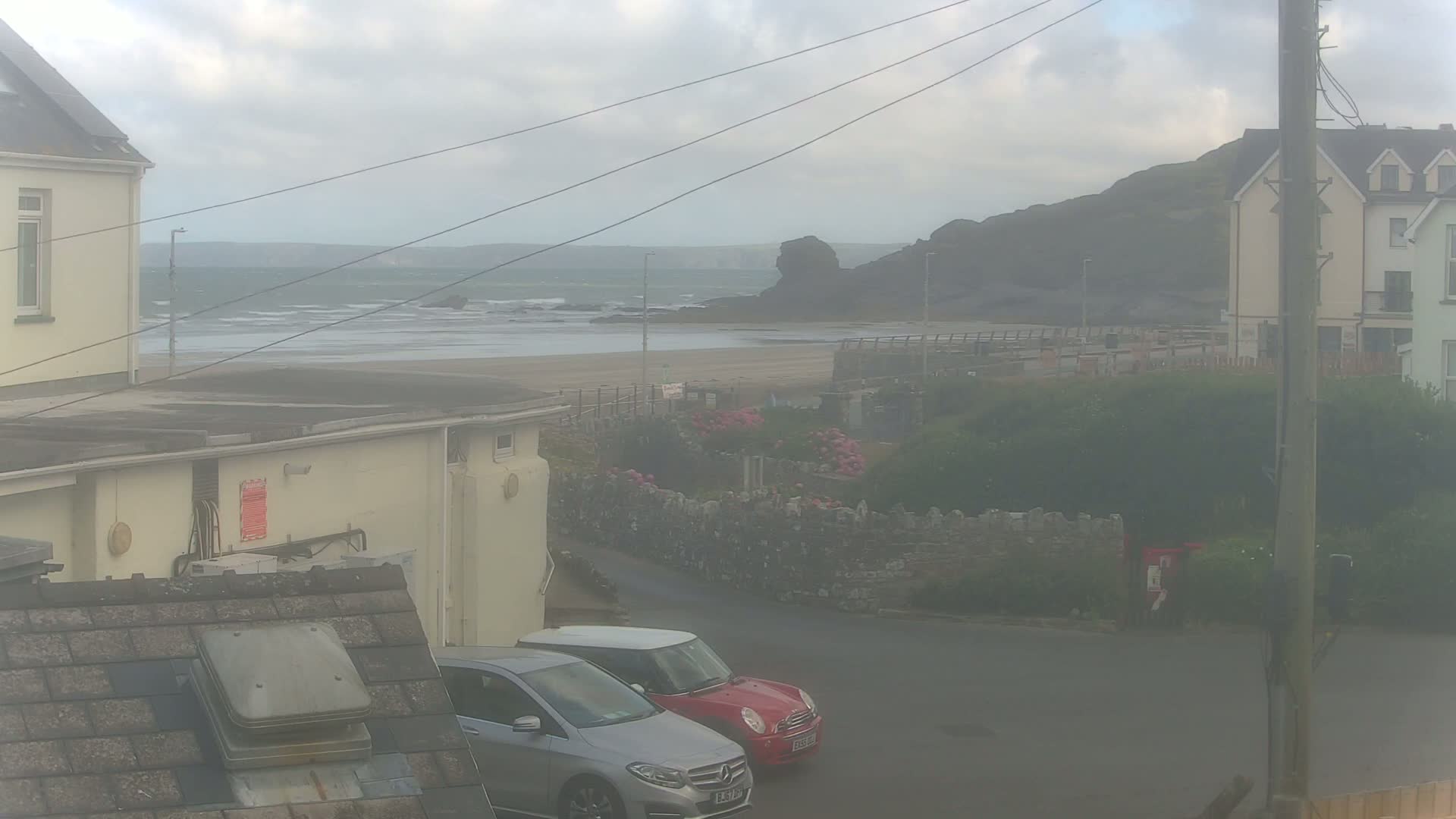 A hazy view of a beach with a calm sea, seen from between two buildings, with two cars parked in a small lot nearby.