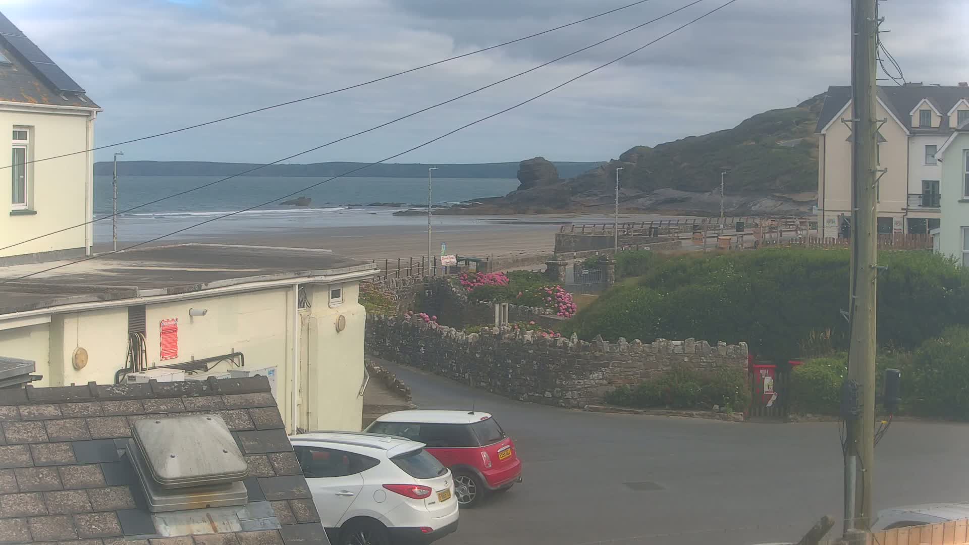 A view from a rooftop shows a sandy beach and calm ocean under an overcast sky, with two cars parked near buildings and a stone wall with pink flowers.