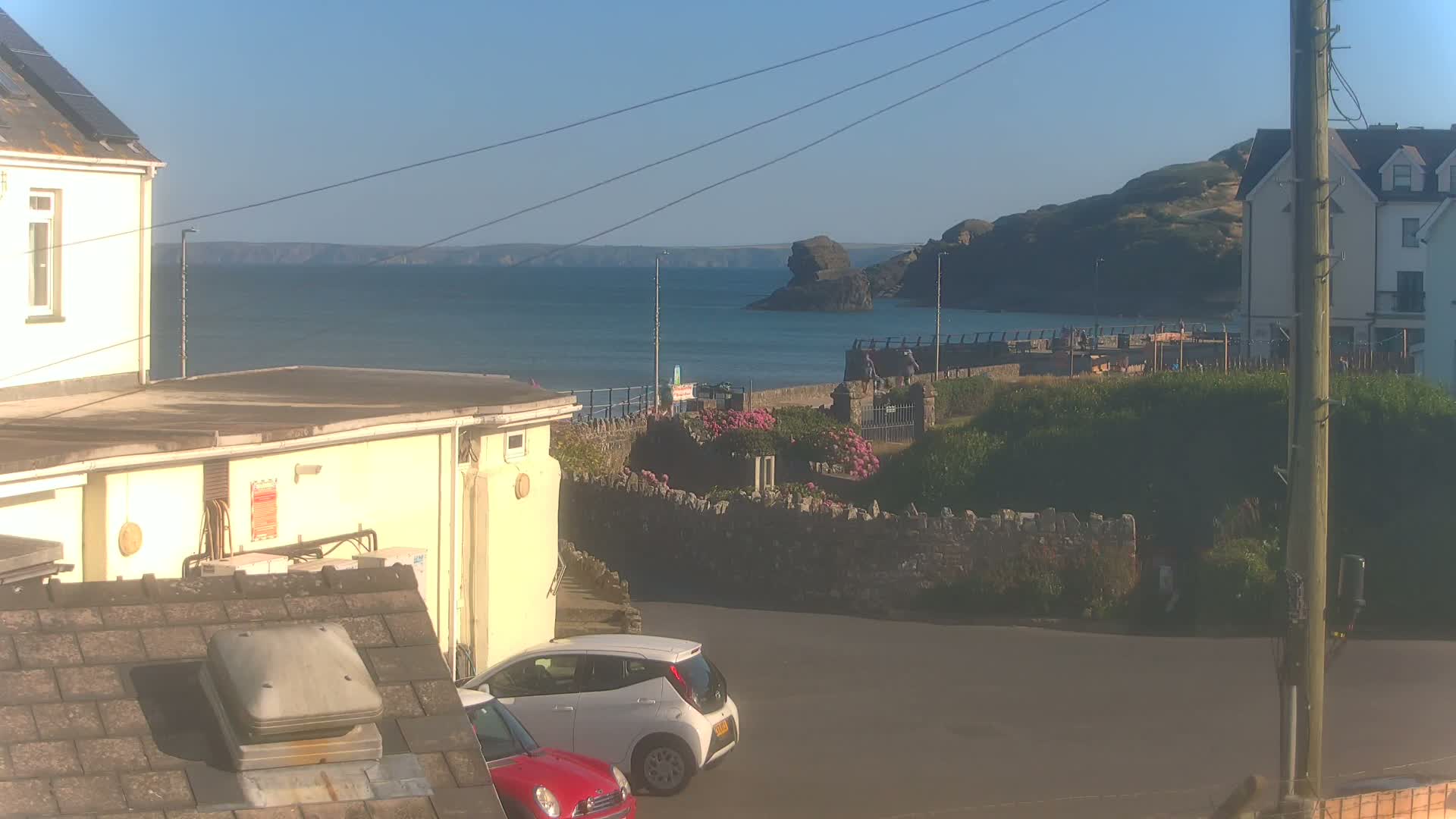 A sunny seaside view shows a calm ocean, a rocky shoreline with buildings and vegetation, and two parked cars in a small parking lot.