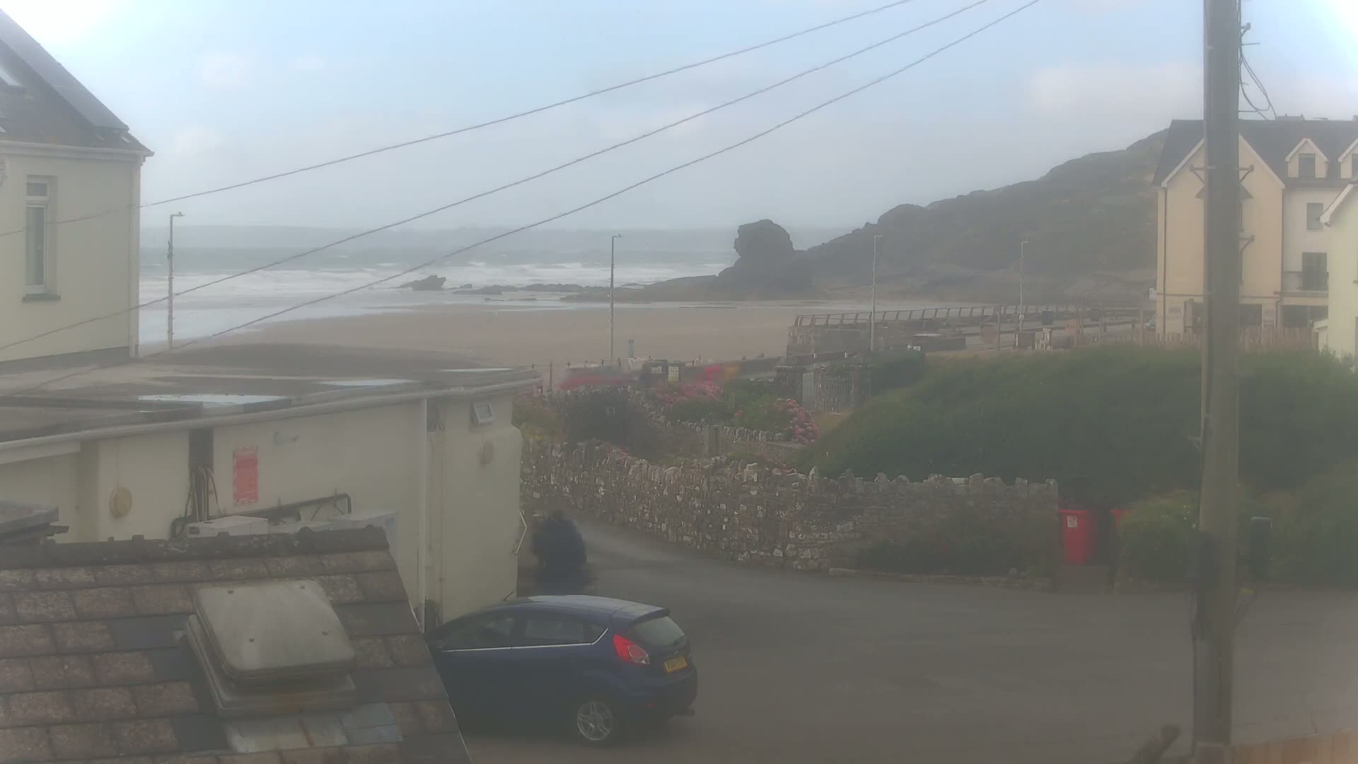 A hazy view of a sandy beach with some small waves, seen from between houses on an overcast day, with a blue car parked on a road in the foreground.
