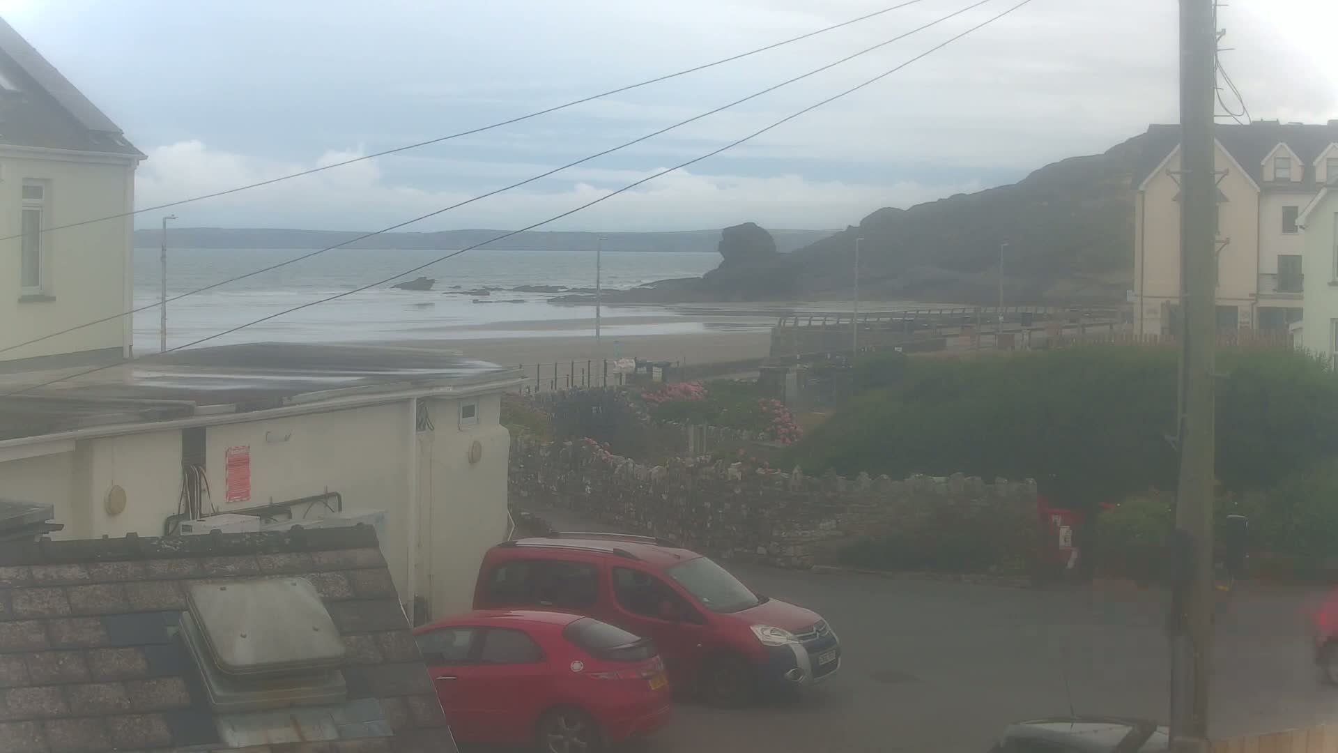 A view from a window shows a hazy, overcast day over a beach with calm water, two red cars parked on a street, and buildings on either side of the street.