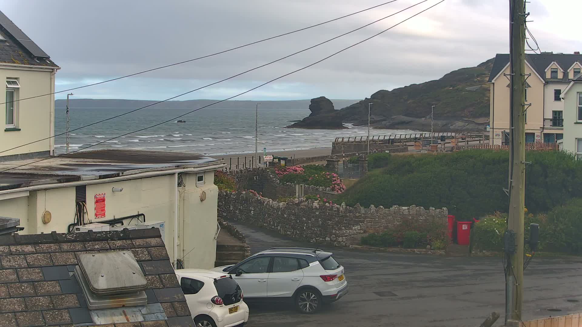 A seaside scene on an overcast day shows a sandy beach with waves, buildings, and two parked cars; a stone wall separates the buildings from the beach and a parking area.