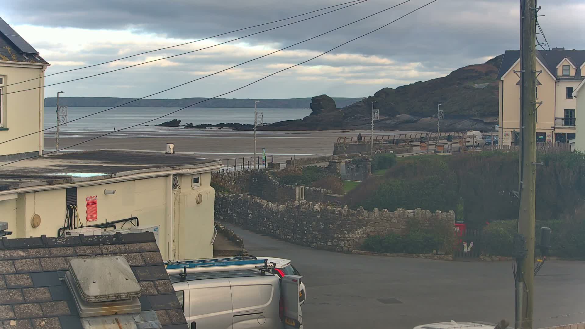 A view of a coastal town on an overcast day shows buildings, a road with a parked van, a stone wall, a promenade with lamp posts, a wide sandy beach, and the sea with rocky formations and distant land under a cloudy sky.