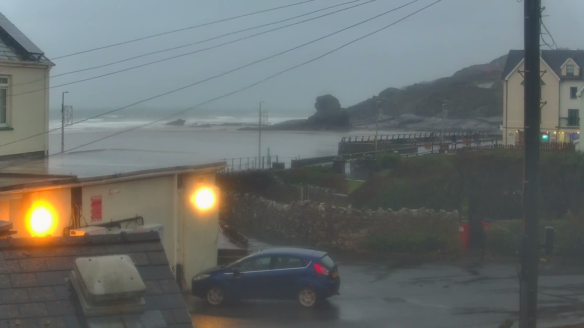 A dark blue car is parked on a wet street in a coastal town with building lights illuminated, facing a turbulent grey sea and a rocky coastline under an overcast and rainy sky.