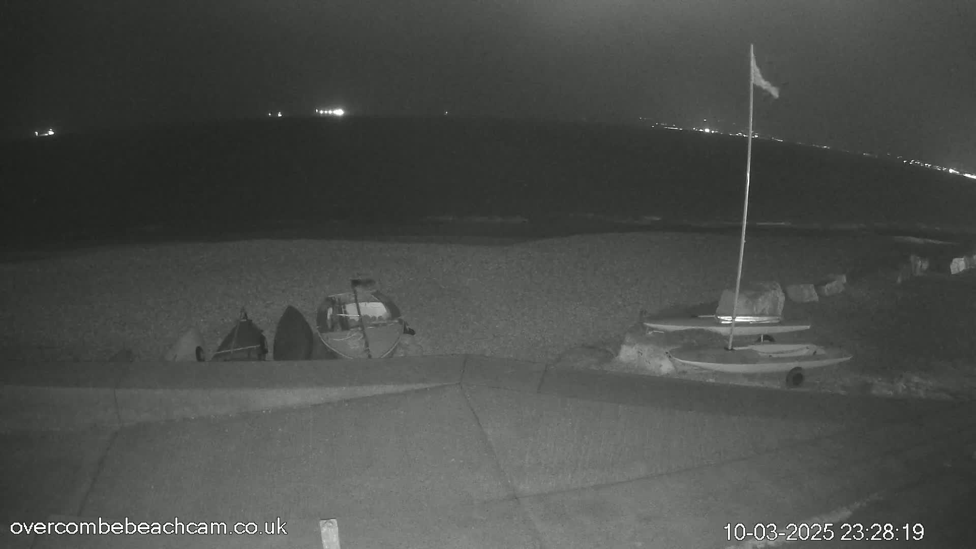 Several small boats are beached on a pebble shore at night under a dark sky with distant lights visible across the water.