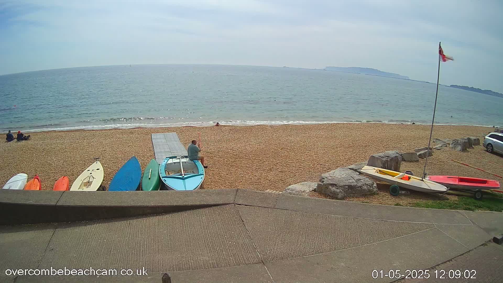 A sandy beach with several small boats and a person sitting near them, overlooks a calm sea under a sunny sky.