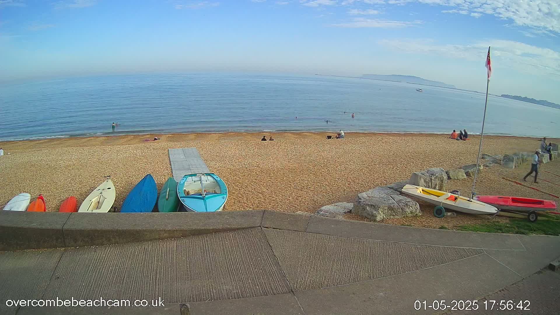 Several small boats are lined up on a pebble beach next to a calm sea under a mostly clear sky, with a few people visible on the beach and in the water.