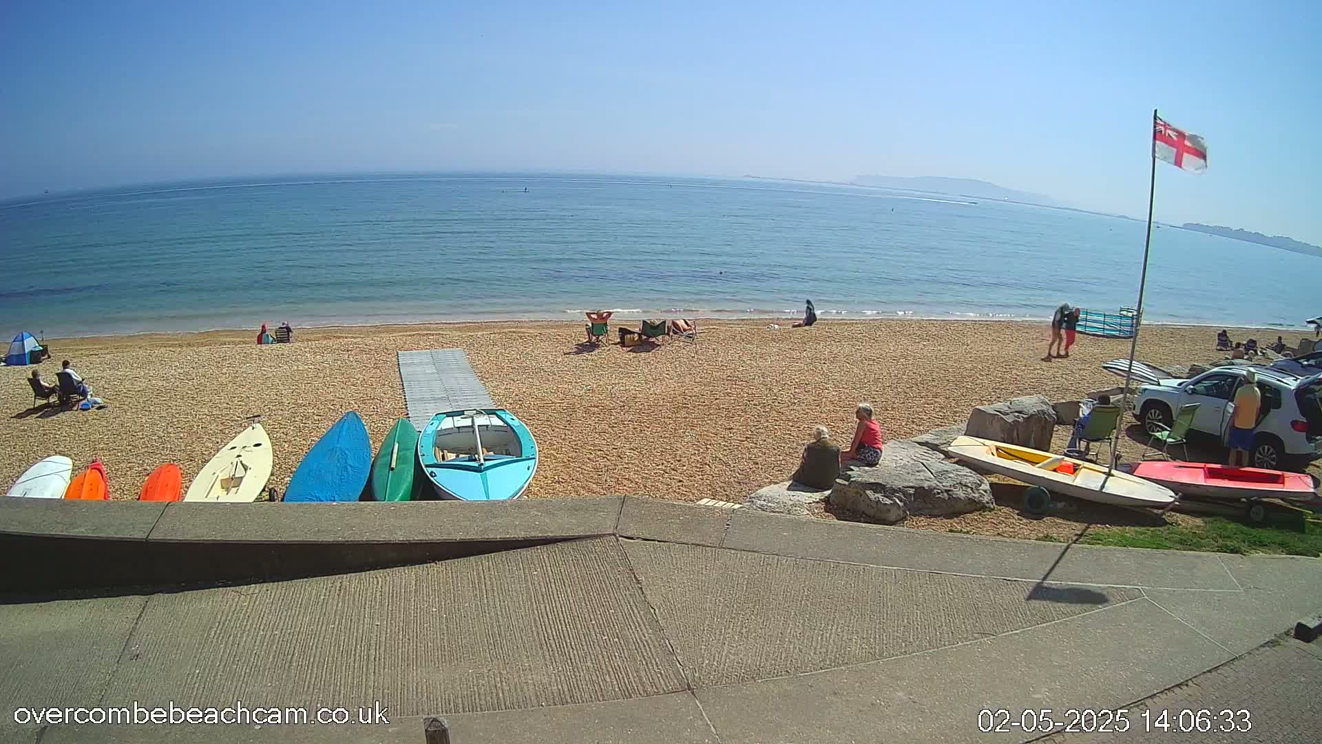 A sunny beach scene shows several people relaxing on the sand near a calm ocean, with small boats and surfboards lined up on the shore.