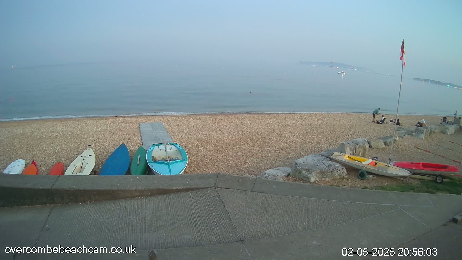 Several small boats are lined up on a pebble beach next to a calm sea under a hazy sky.