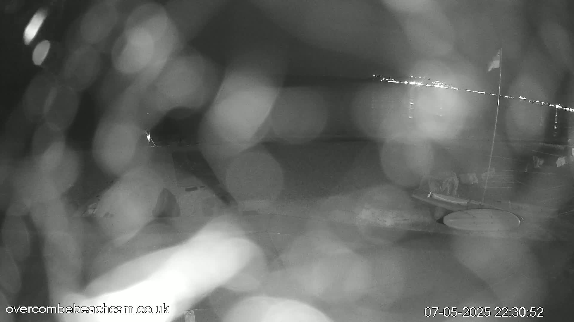 A blurry nighttime image shows a dark beach with a distant line of lights, possibly from a town, and rain or moisture obscuring the view.