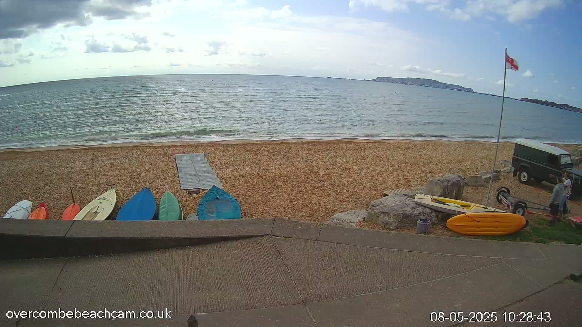 A sandy beach with several small boats and a Land Rover parked nearby, overlooks a calm sea under a partly cloudy sky.