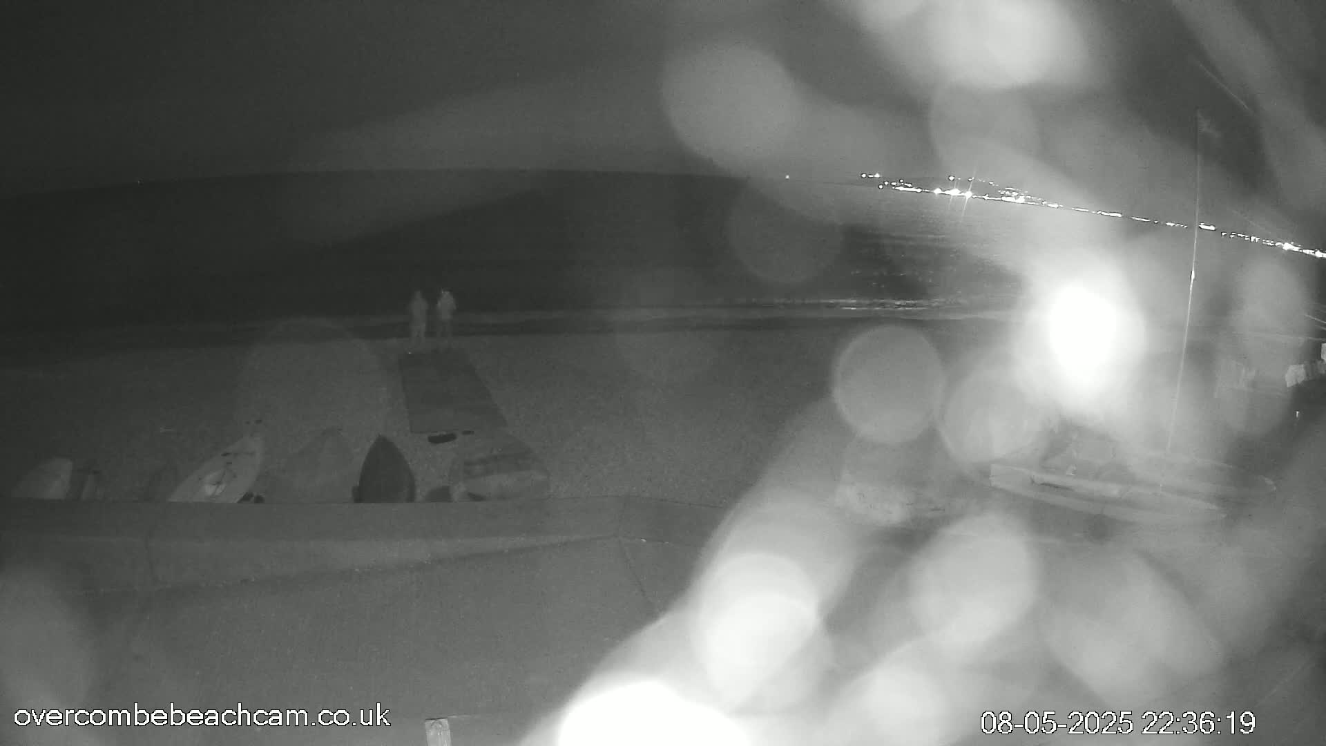 A nighttime, blurry view of a beach shows two figures standing near several items on the sand, with a calm sea and distant lights visible in the background.