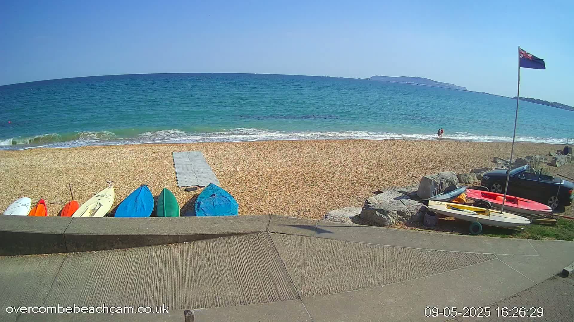 Several small boats are lined up on a pebble beach next to a calm, teal ocean under a clear blue sky.
